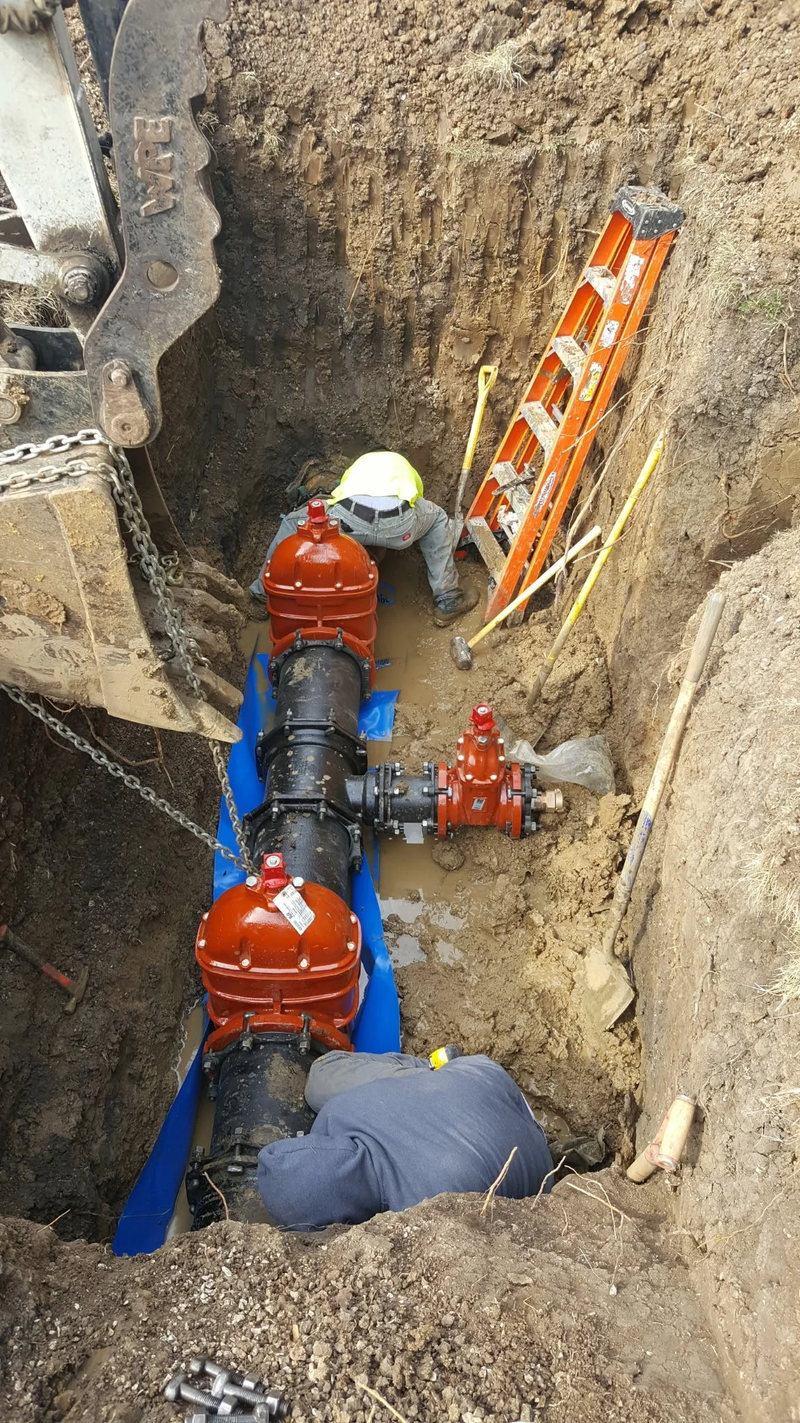 Two workers are installing or repairing large underground pipes in a deep trench with soil walls, using machinery, ladders, and tools.