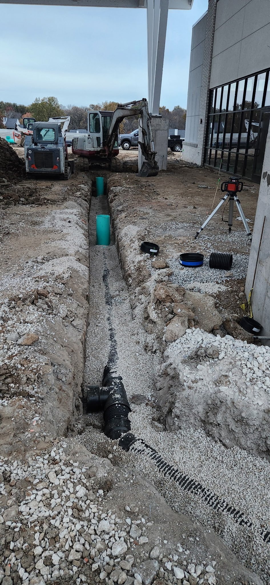 Construction site with trench for underground piping, two green pipes installed, construction equipment, and tools in front of a modern building.