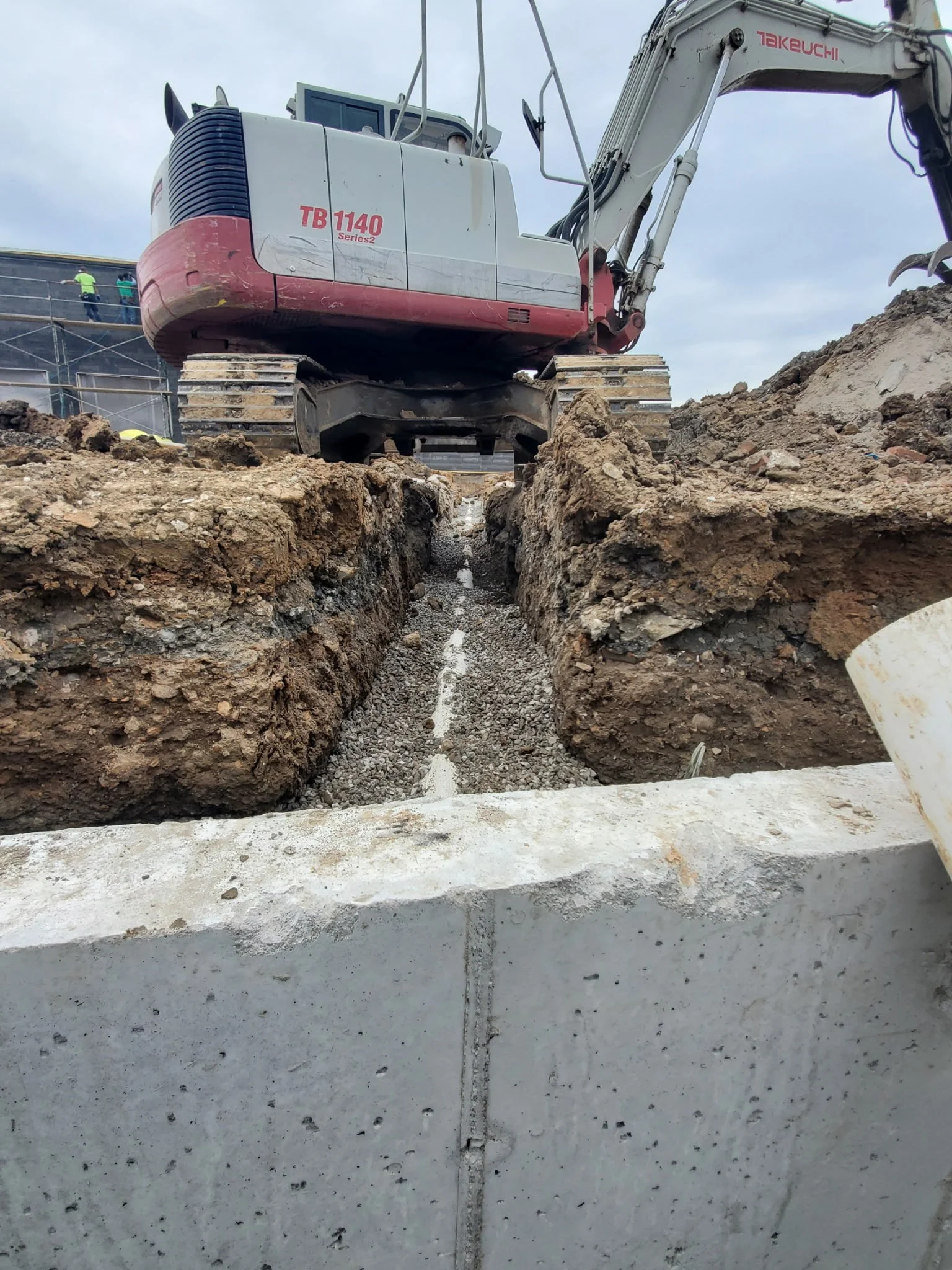 View of a construction site with a large pink and white excavator labeled 'Takeuchi,' positioned over a deep trench with dirt and gravel inside. The background shows a cloudy sky and some construction workers on scaffolding.