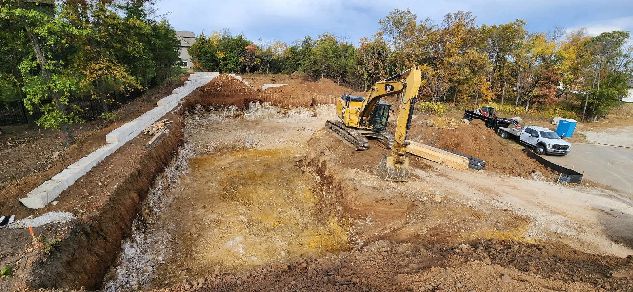 Construction site with an excavator working on the dirt, concrete blocks along the edge, trucks, a portable toilet, and trees with autumn foliage in the background.