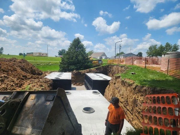 Construction site with large concrete drainage structures being installed in a deep trench, with a worker in an orange shirt and a baseball cap supervising, under a partly cloudy sky.