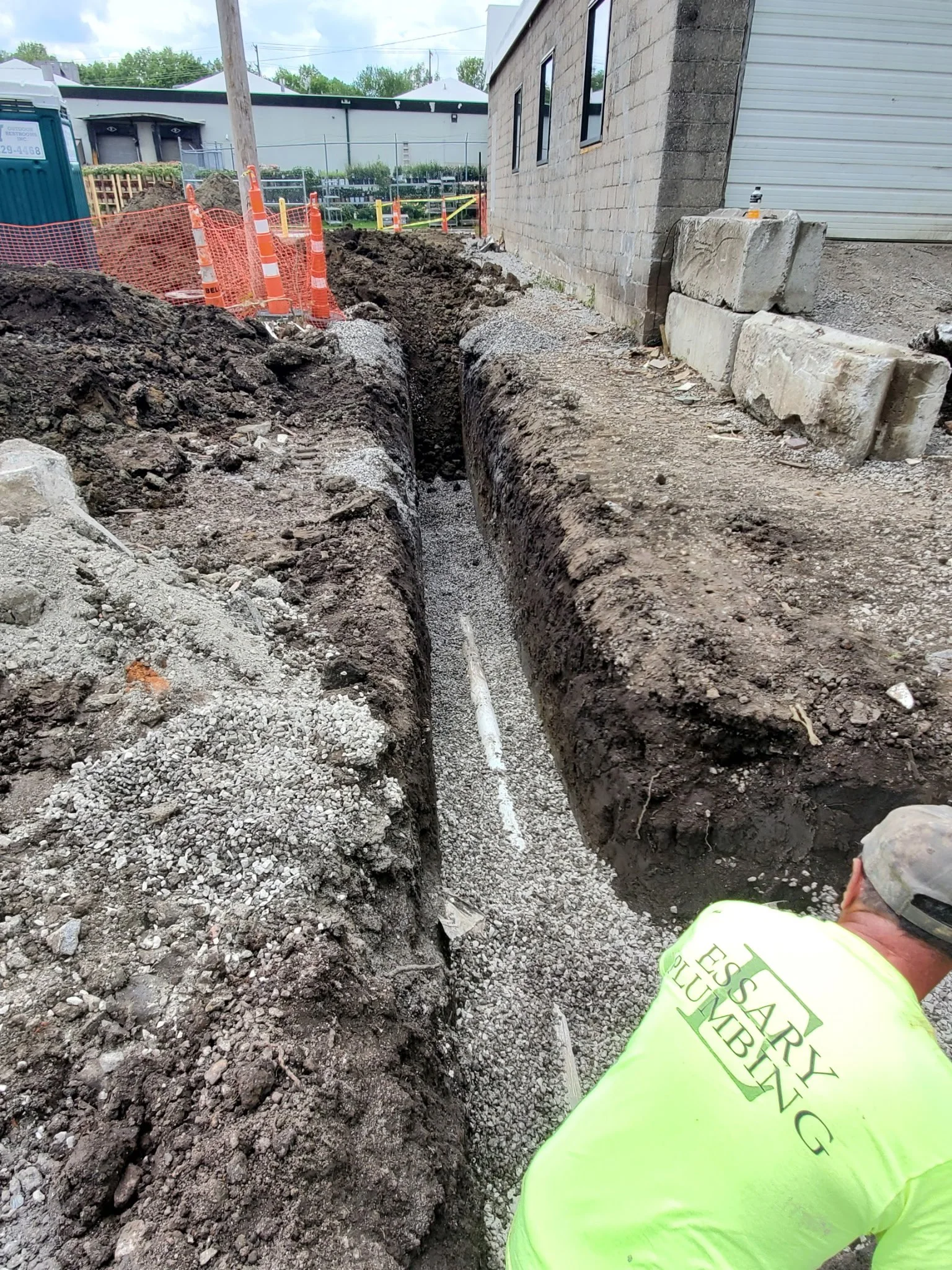 Construction worker in a yellow high-visibility shirt working in a trench for underground utility installation or repair on a city street, with construction barriers and building in the background.