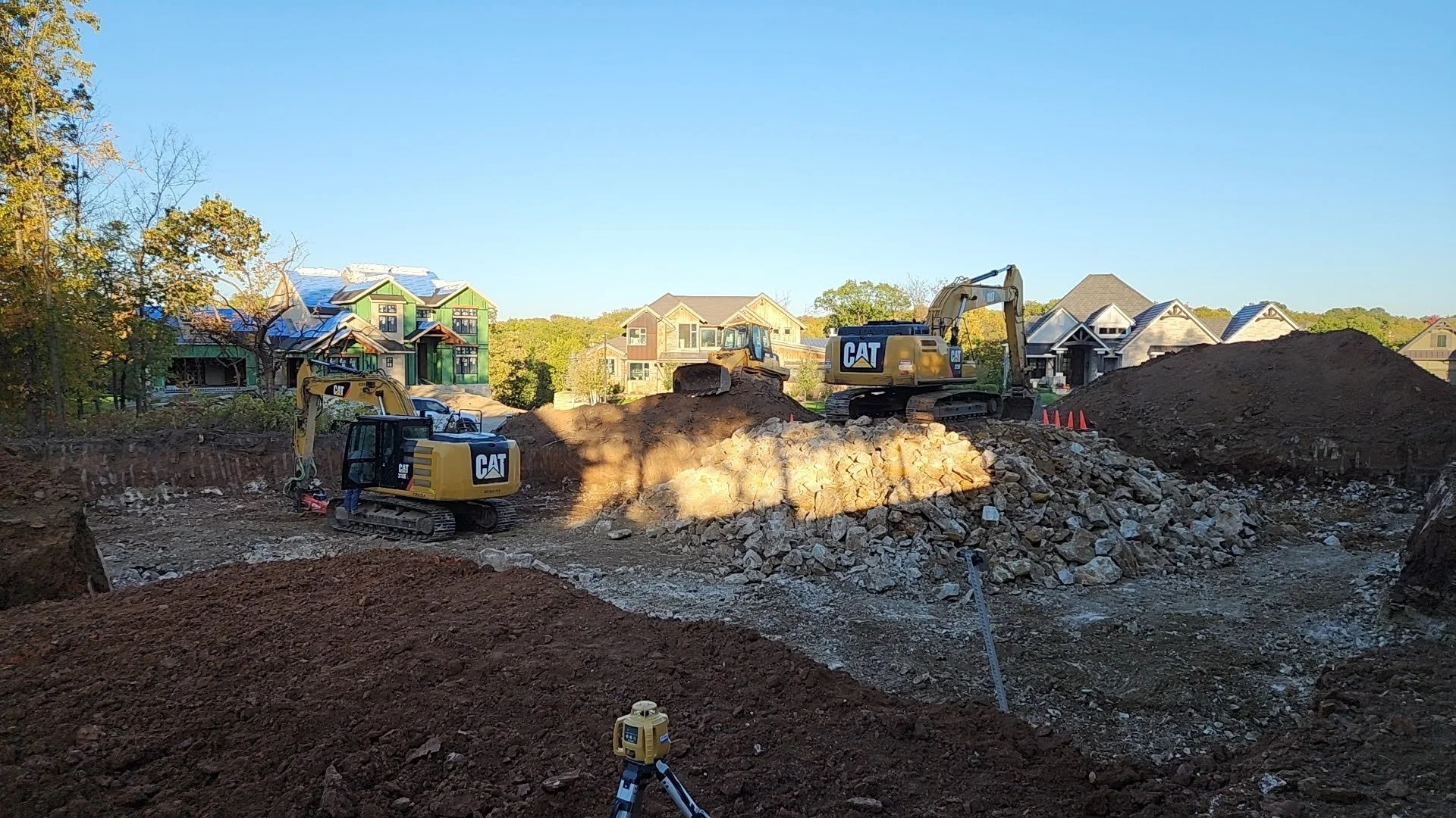 Construction site with three CAT excavators working on digging and moving earth, surrounded by dirt piles, with residential houses in the background under a clear blue sky.