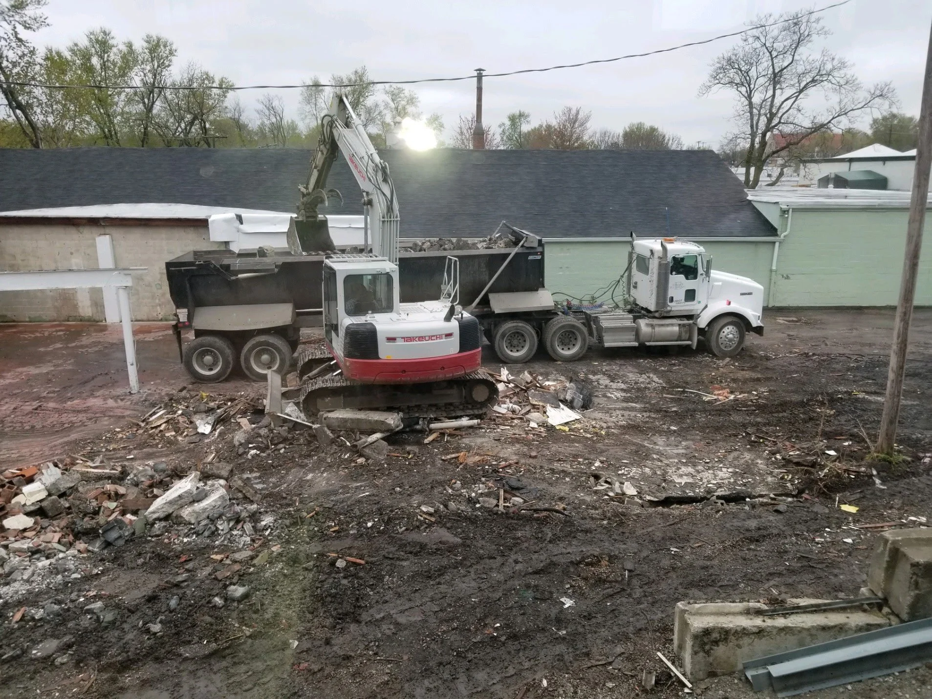 Construction site with a small excavator loading debris into a dump truck on a dirt ground with a building in the background.