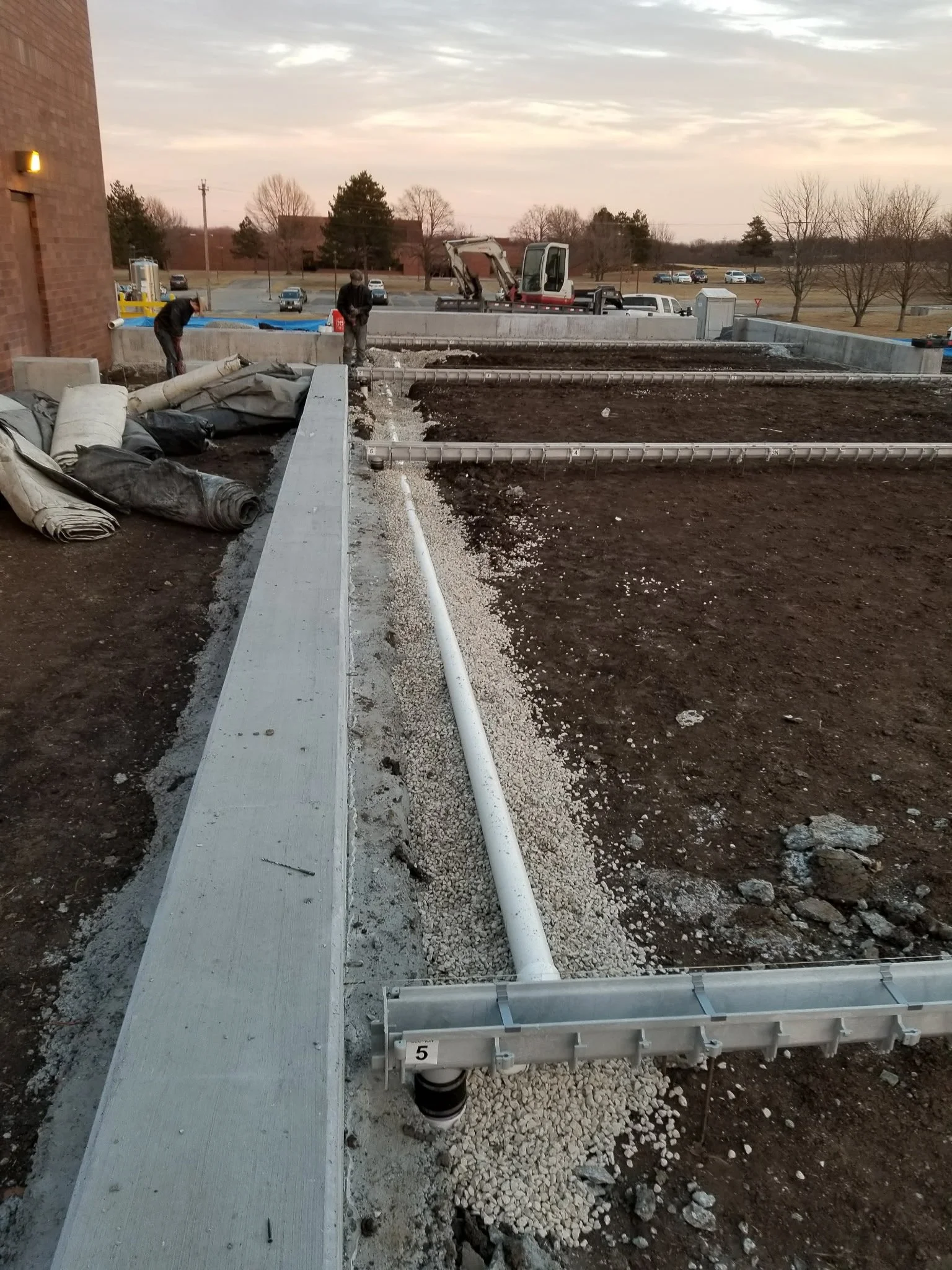 Construction workers installing pipes and gravel on a building rooftop with machinery and parked cars in the background at sunset.