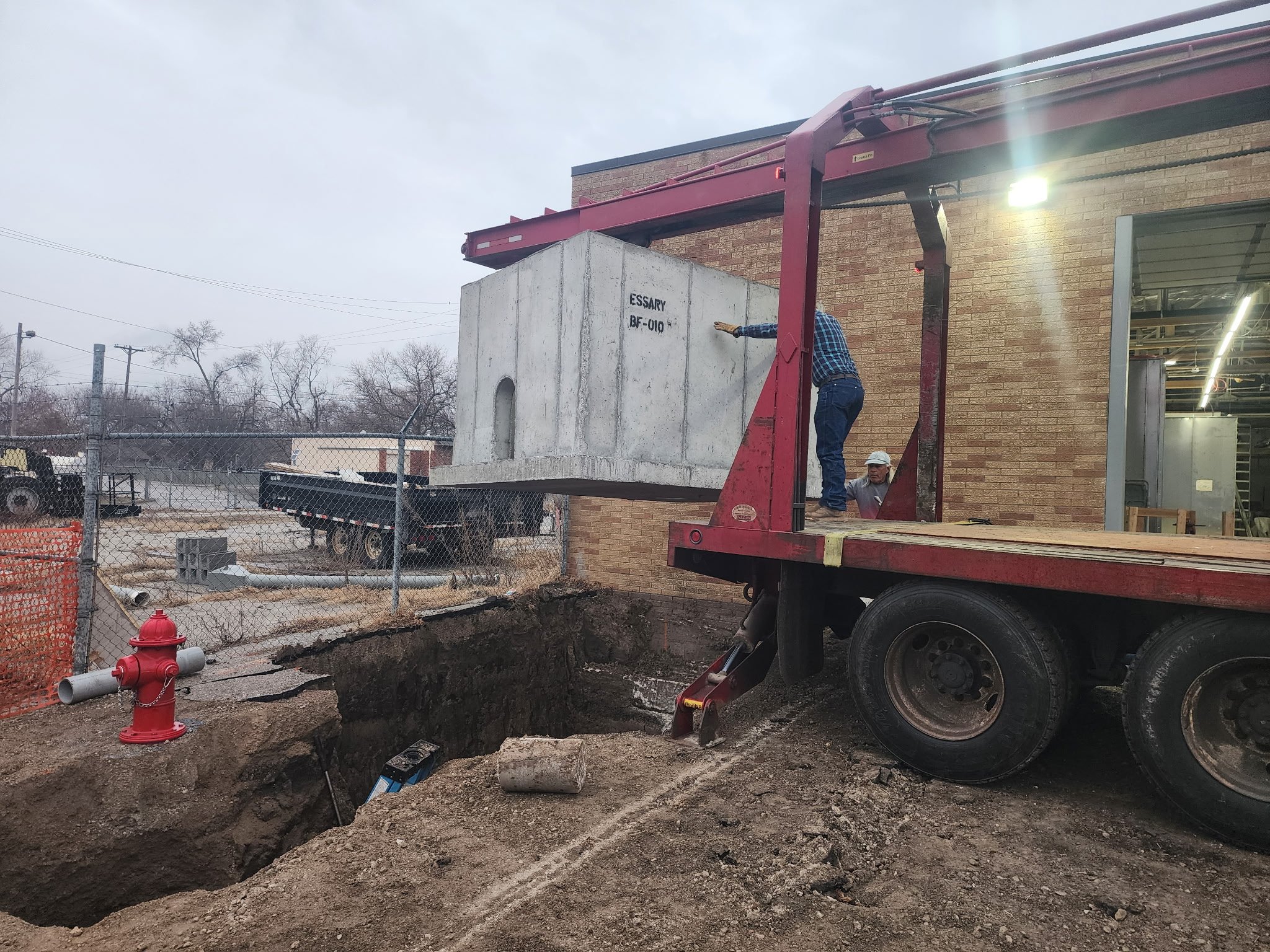Workers using a crane to lift a large concrete block at a construction site outside a brick building, with construction equipment and vehicles in the background.