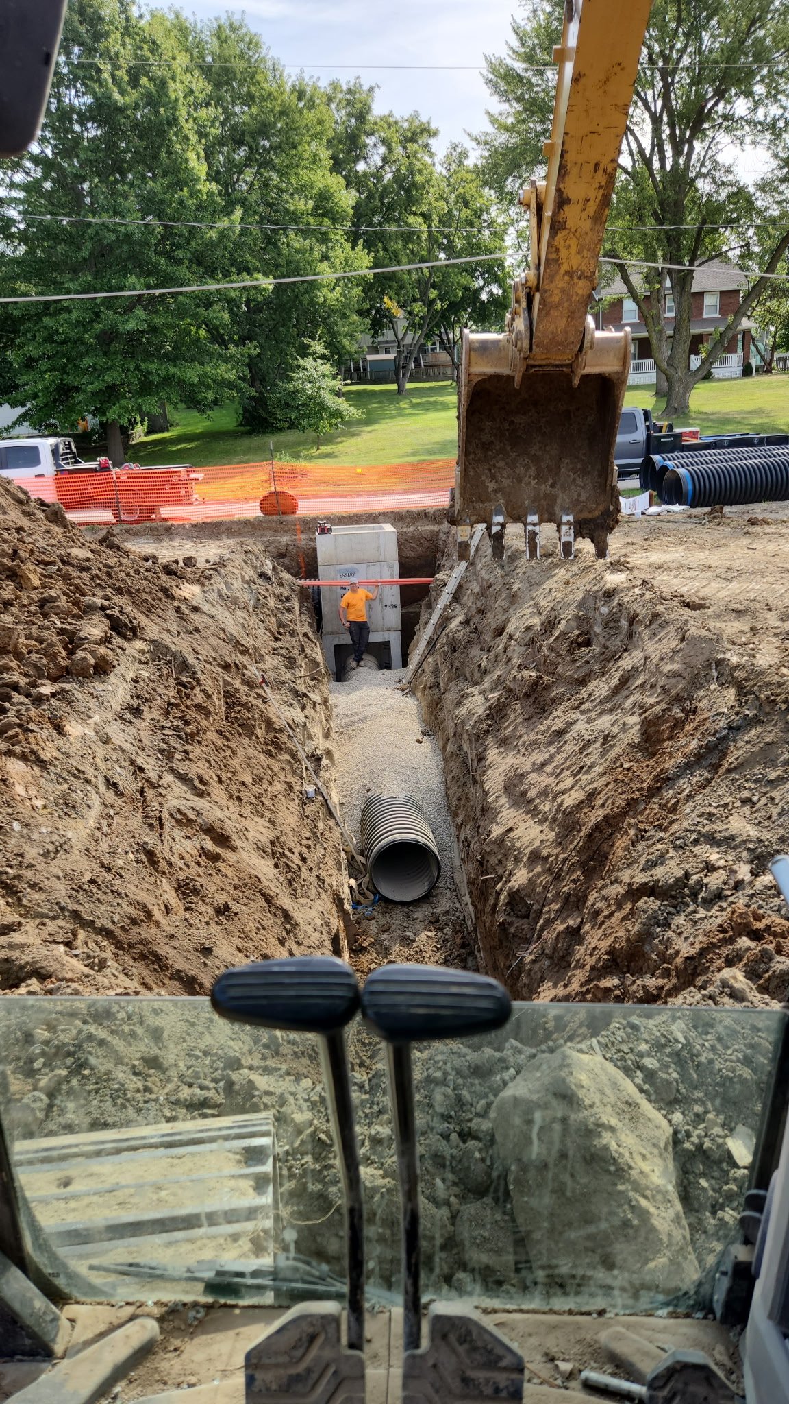 Construction site with excavator digging a trench for underground piping, a large pipe laid in the trench, and a construction worker near a utility box in the background.