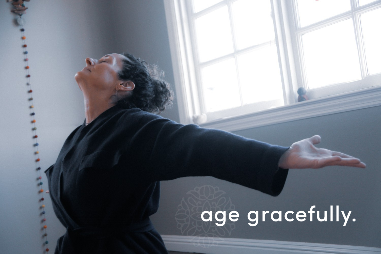 A woman with curly hair and earrings practicing yoga or stretching indoors, with sunlight coming through a window, and the phrase "age gracefully" written on the image.