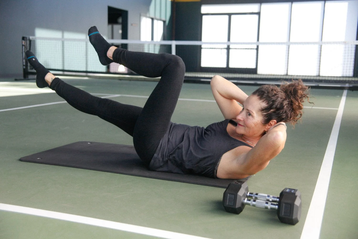 A woman doing abdominal exercises on a yoga mat in a gym, with a dumbbell nearby.