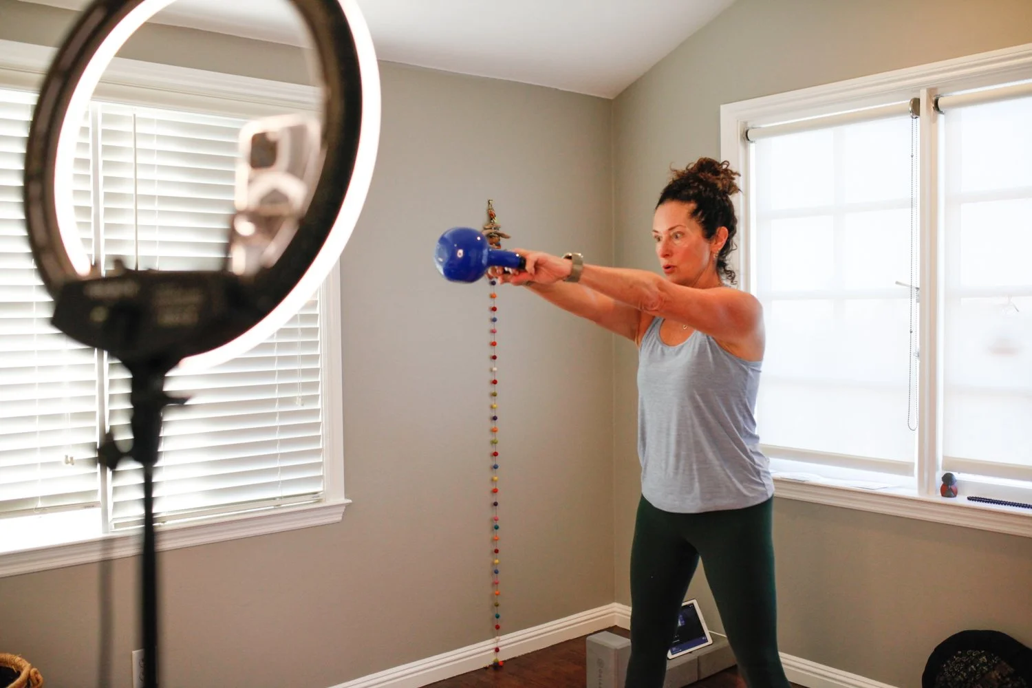 Woman doing an exercise with a blue kettlebell in a bright room, with a ring light and camera setup for recording or streaming.