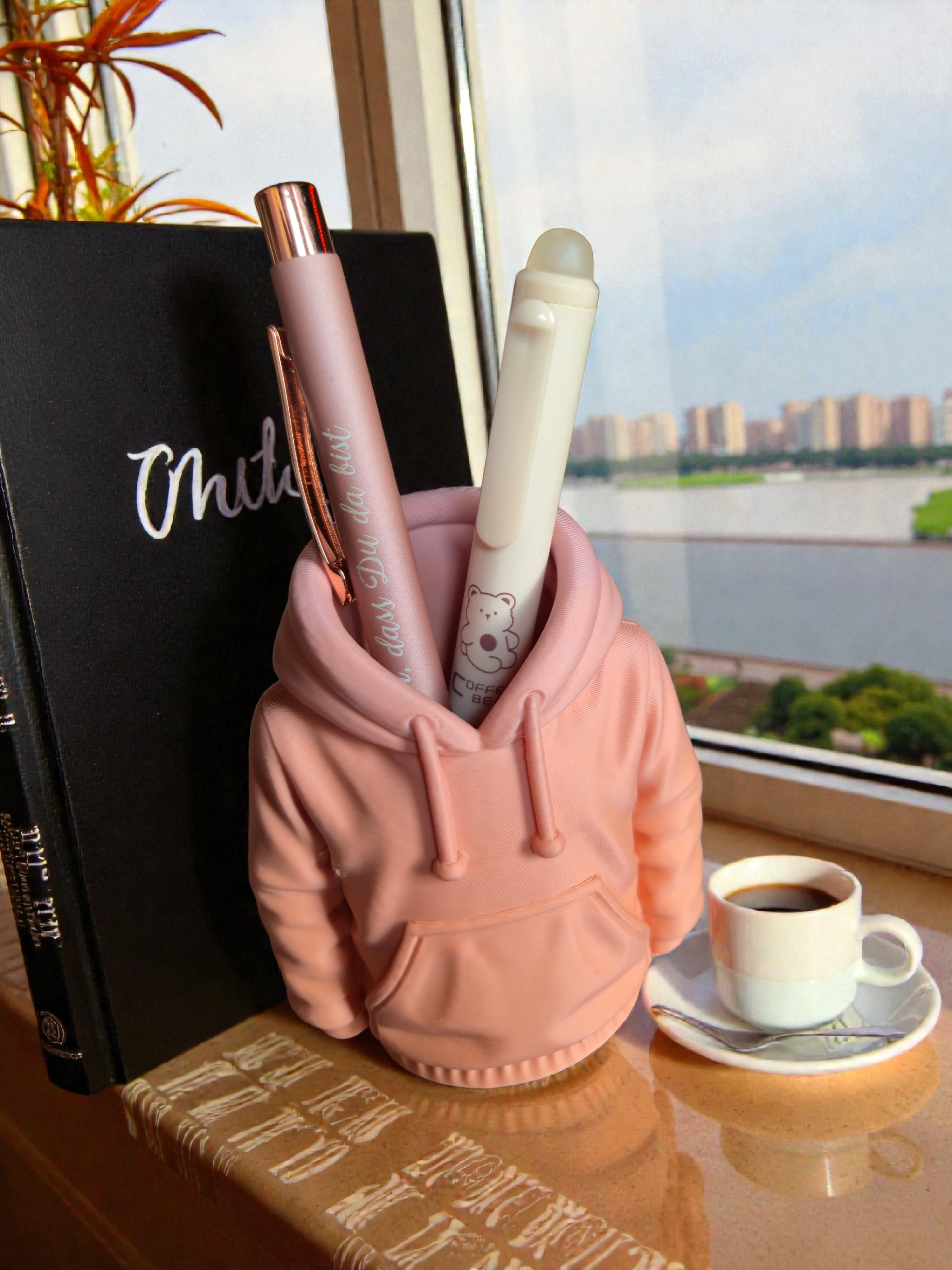 Ein Tassen Kaffee auf einem weißen Teller neben einem Besteck liegt auf einem Tisch, in einer Ecke liegt eine kleine pinke Hoodie mit Stifte darin, vor einem Fenster mit Stadt- und Flussblick im Hintergrund.