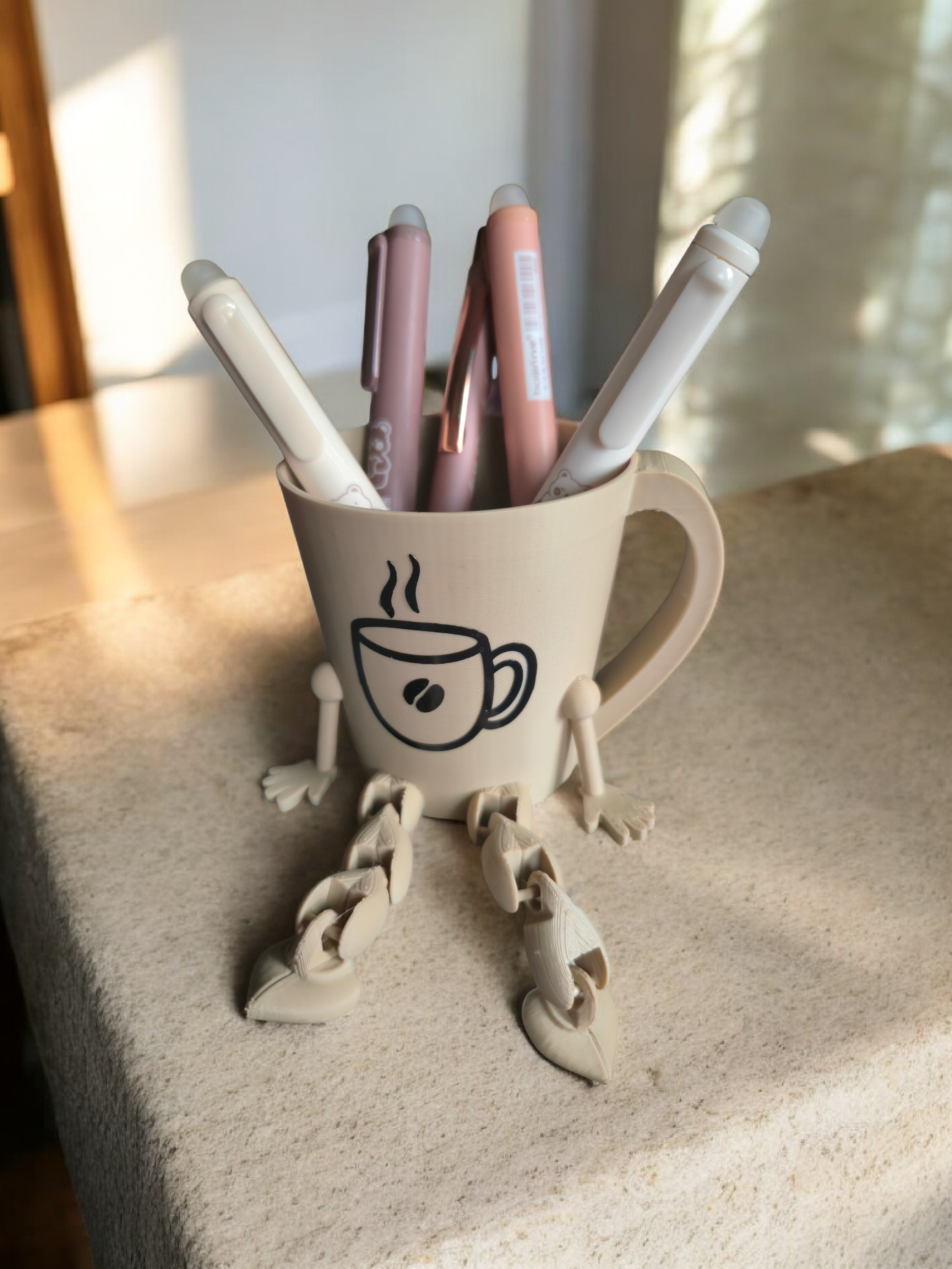 Kaffeetasse mit Fransen und Armen, in der Kaffeetasse sind Stifte, auf einem Tisch.