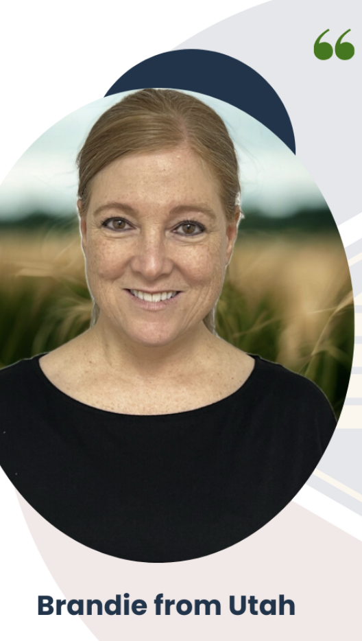 A smiling woman with red hair and freckles, wearing a black shirt, standing outdoors in a wheat field with a blurred green and blue background.