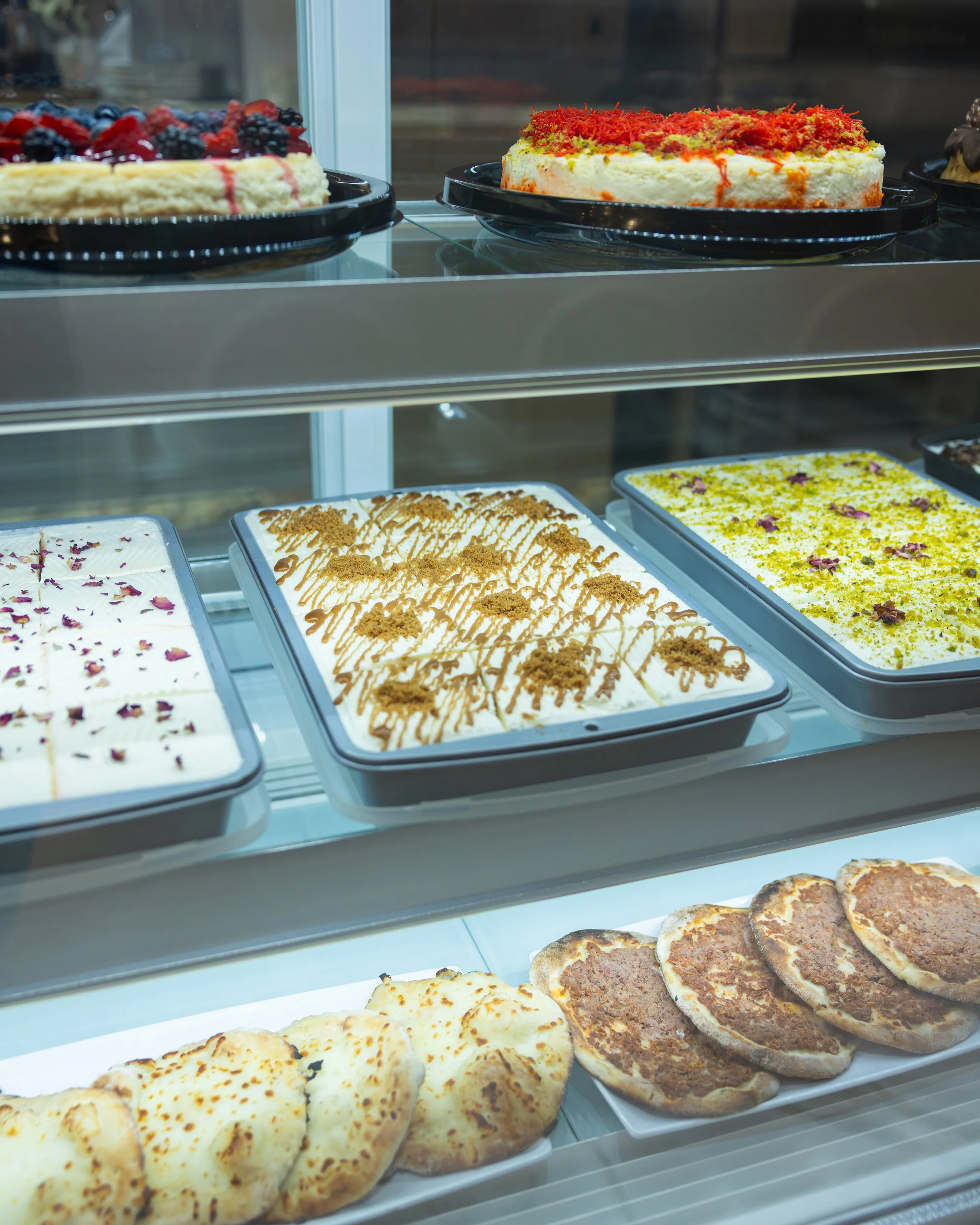 Showcase of assorted desserts in a glass display case, including cheesecakes topped with berries, drizzled caramel, and pistachios, and various types of flatbreads at the bottom.