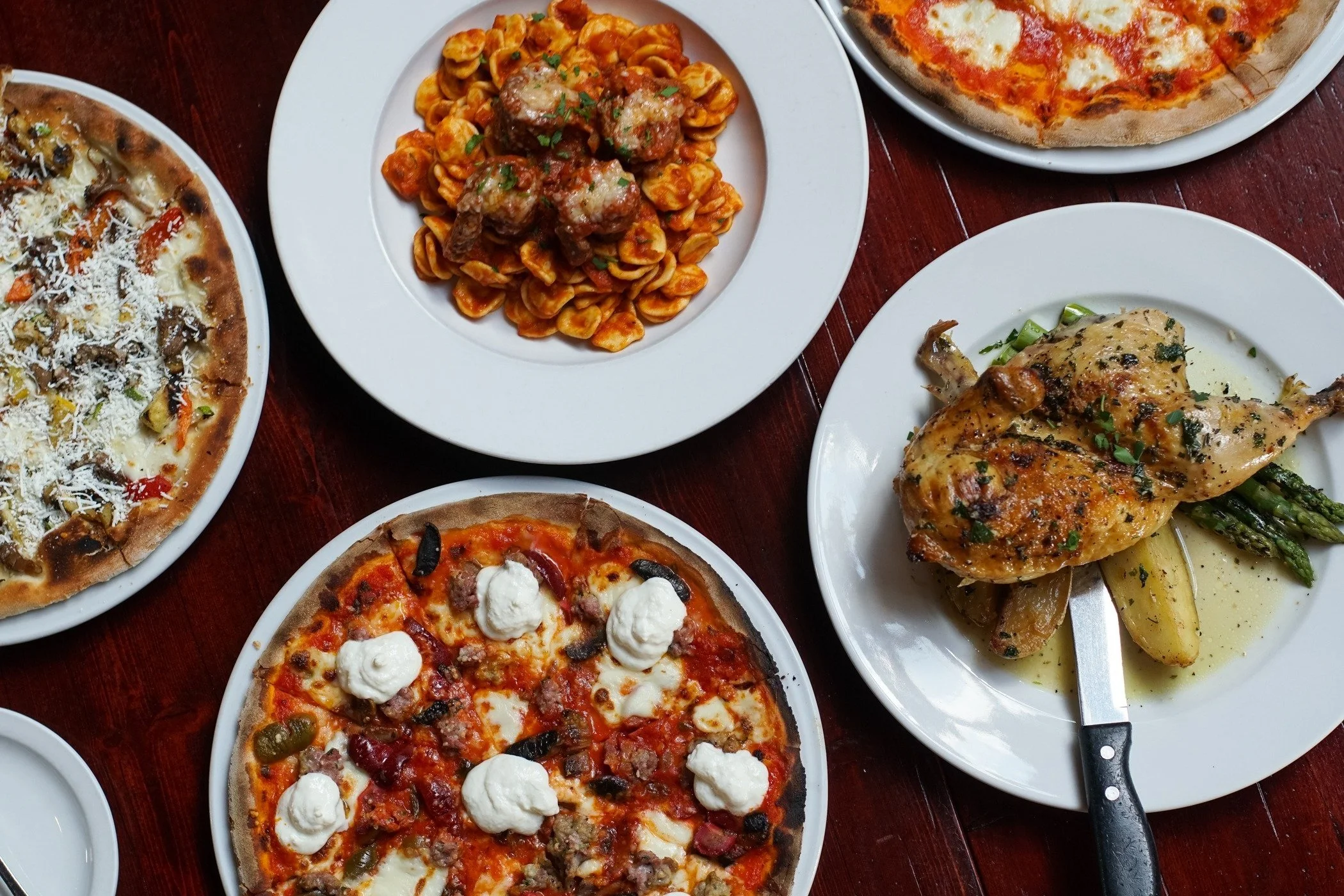 Assorted Italian dishes on white plates, including pizza with cheese and toppings, pasta with meatballs, baked pizza with cheese and herbs, and roasted chicken with potatoes and asparagus, all on a dark wooden table.