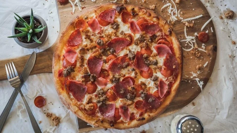 Pepperoni and sausage pizza on a wooden serving board with some scattered toppings, a potted succulent plant, and eating utensils nearby.