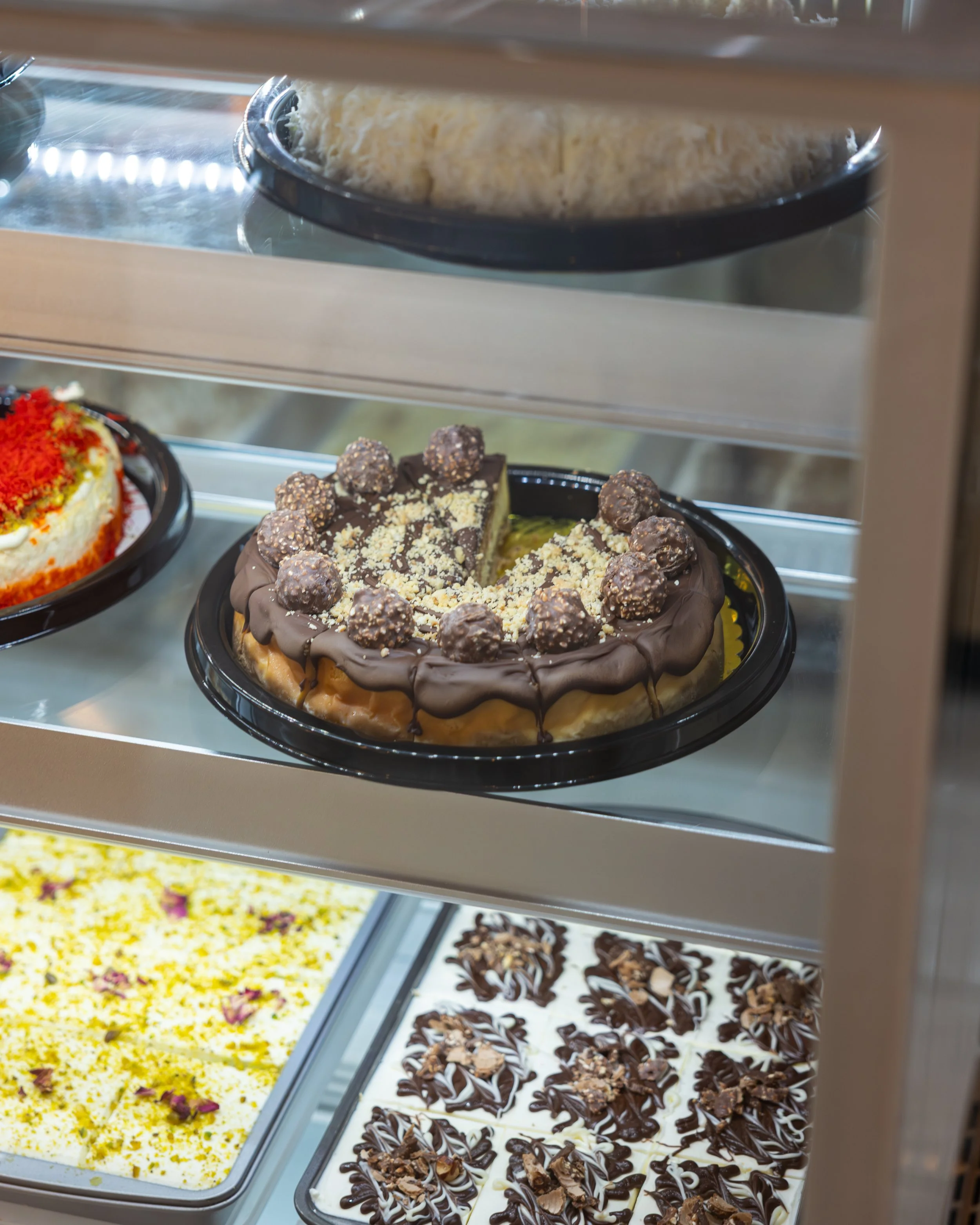 Display case with a chocolate cake topped with Ferrero Rocher chocolates and crumbled toppings, and other desserts below it including a yellow cake with flower petals and a tray of decorated chocolate squares.