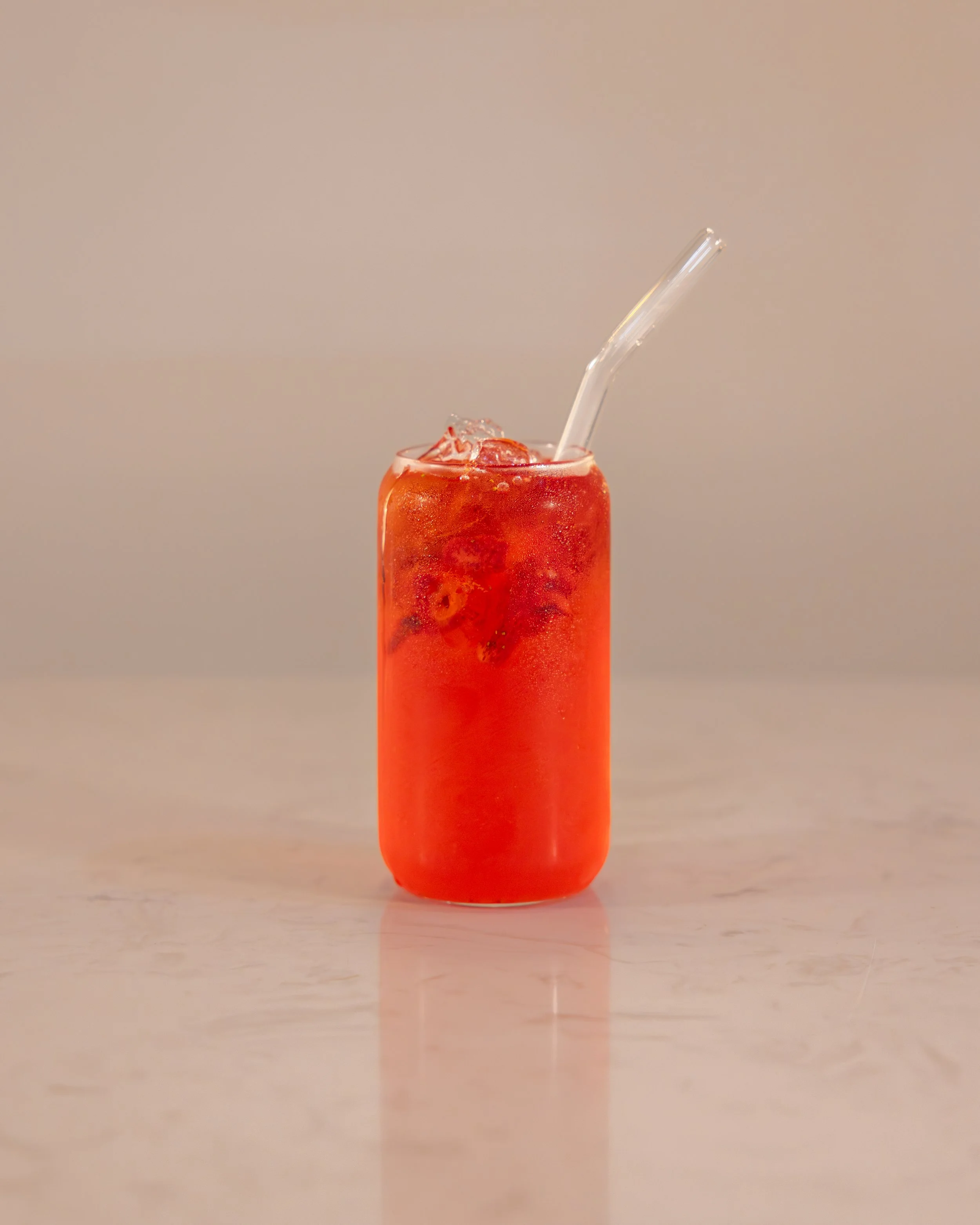 A tall glass filled with a red beverage and ice, with a clear straw, placed on a light-colored surface against a plain background.