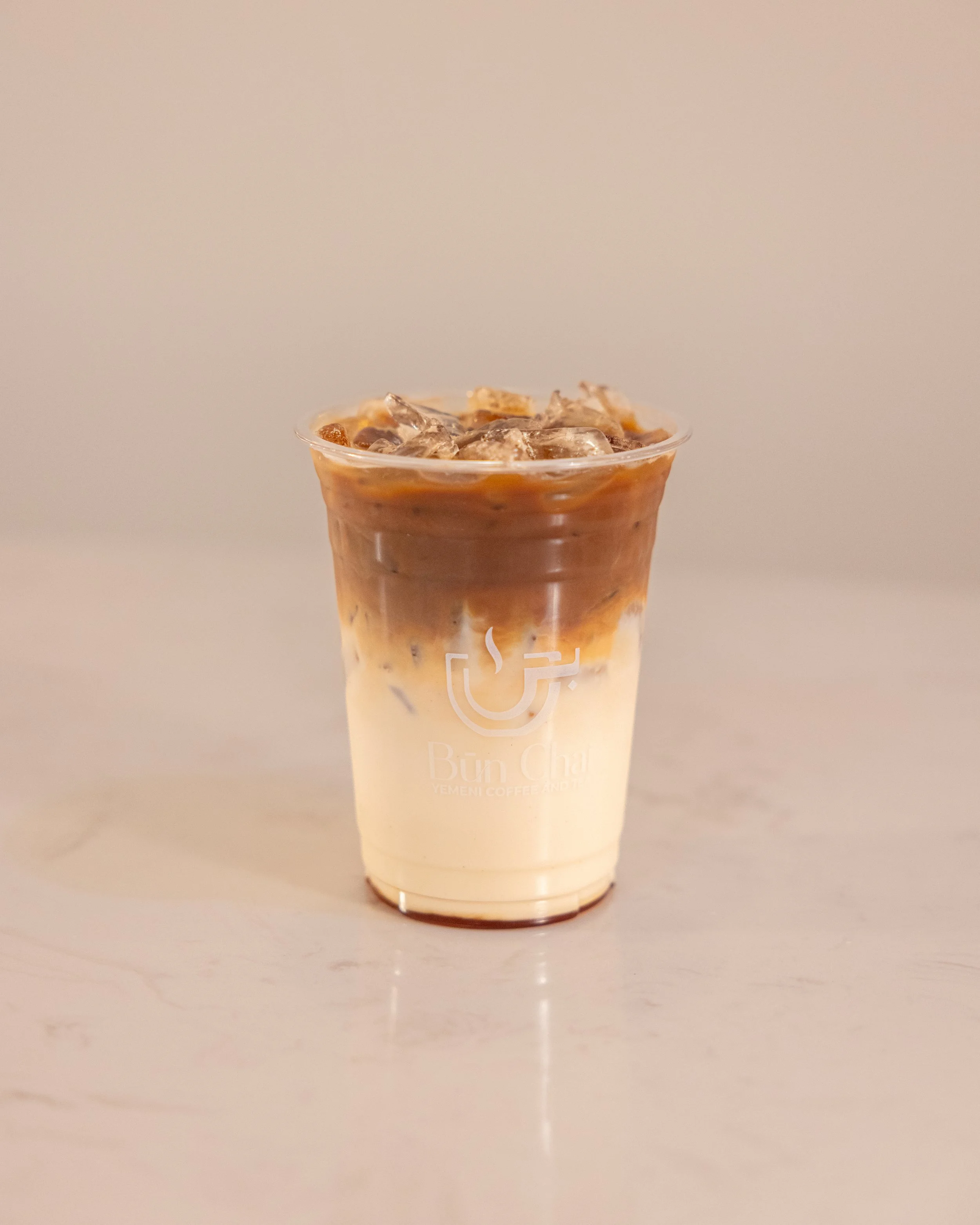 A plastic cup of iced coffee with layers of coffee and milk topped with ice cubes, on a light-colored surface.