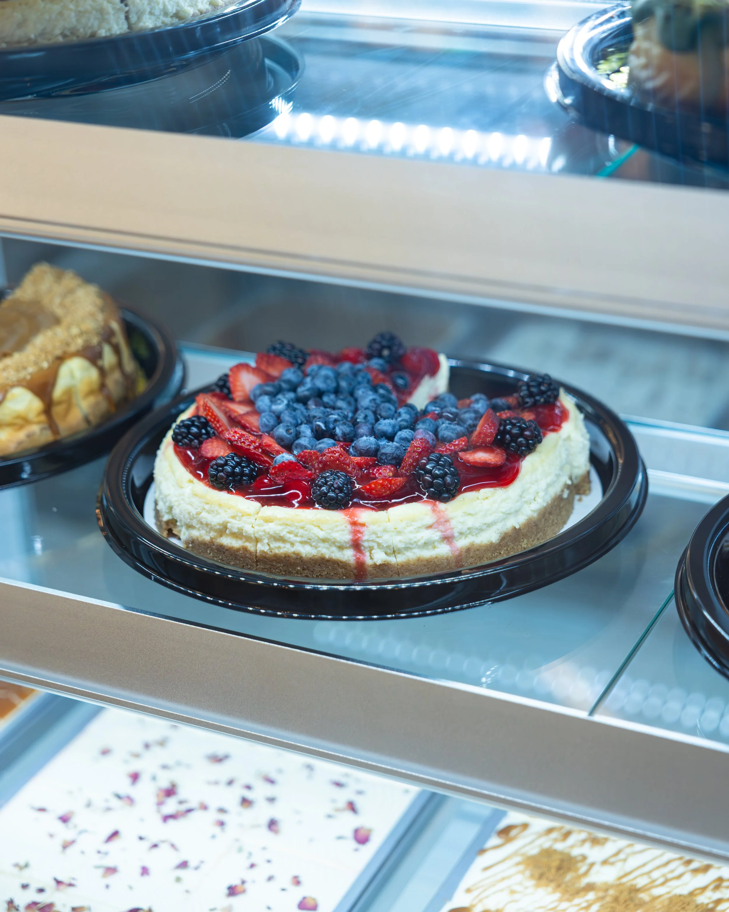 A cheesecake topped with mixed berries including blueberries, strawberries, and blackberries inside a glass display case.