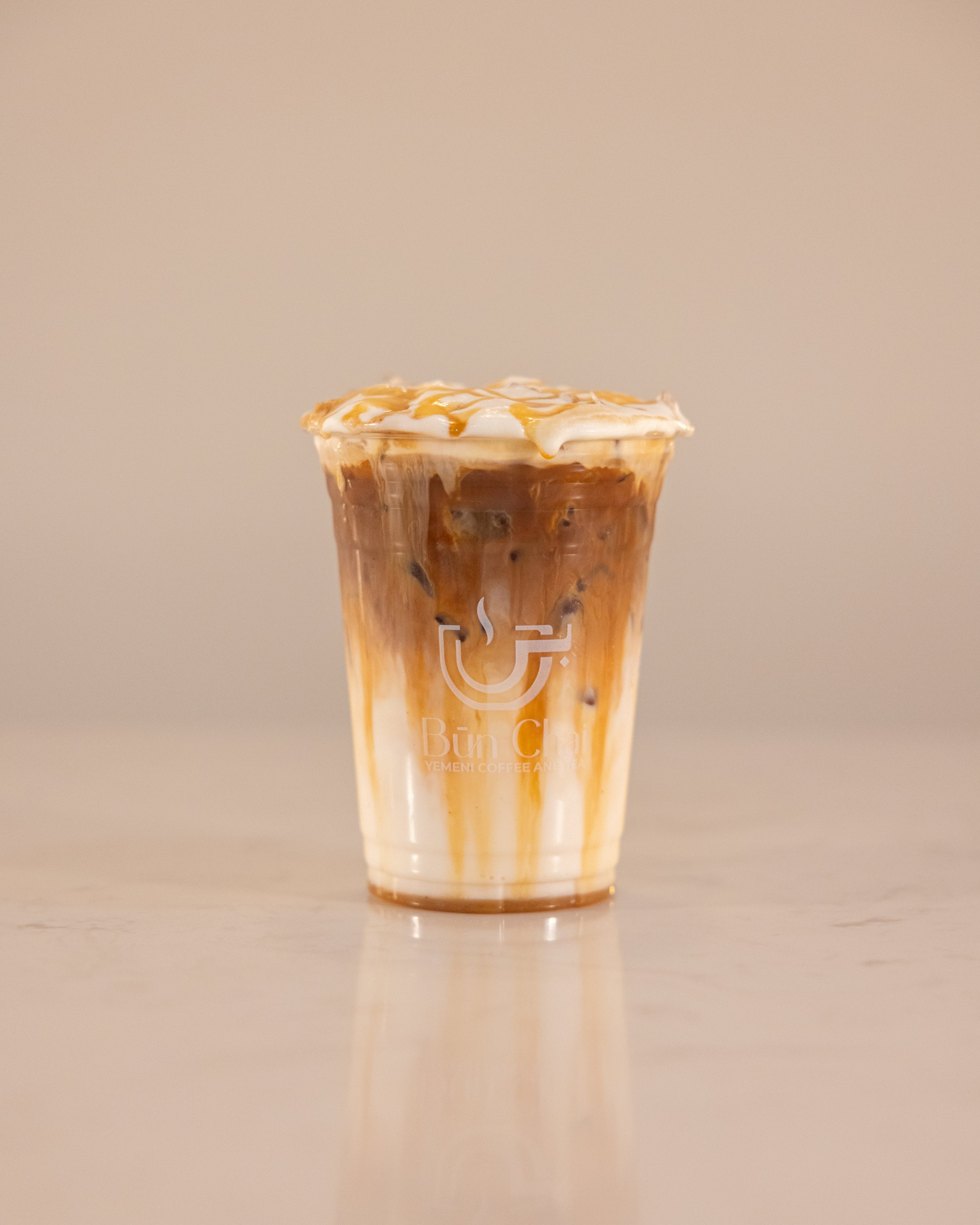 Iced coffee drink with caramel topping in a clear plastic cup with a Bun Cafe logo on it.