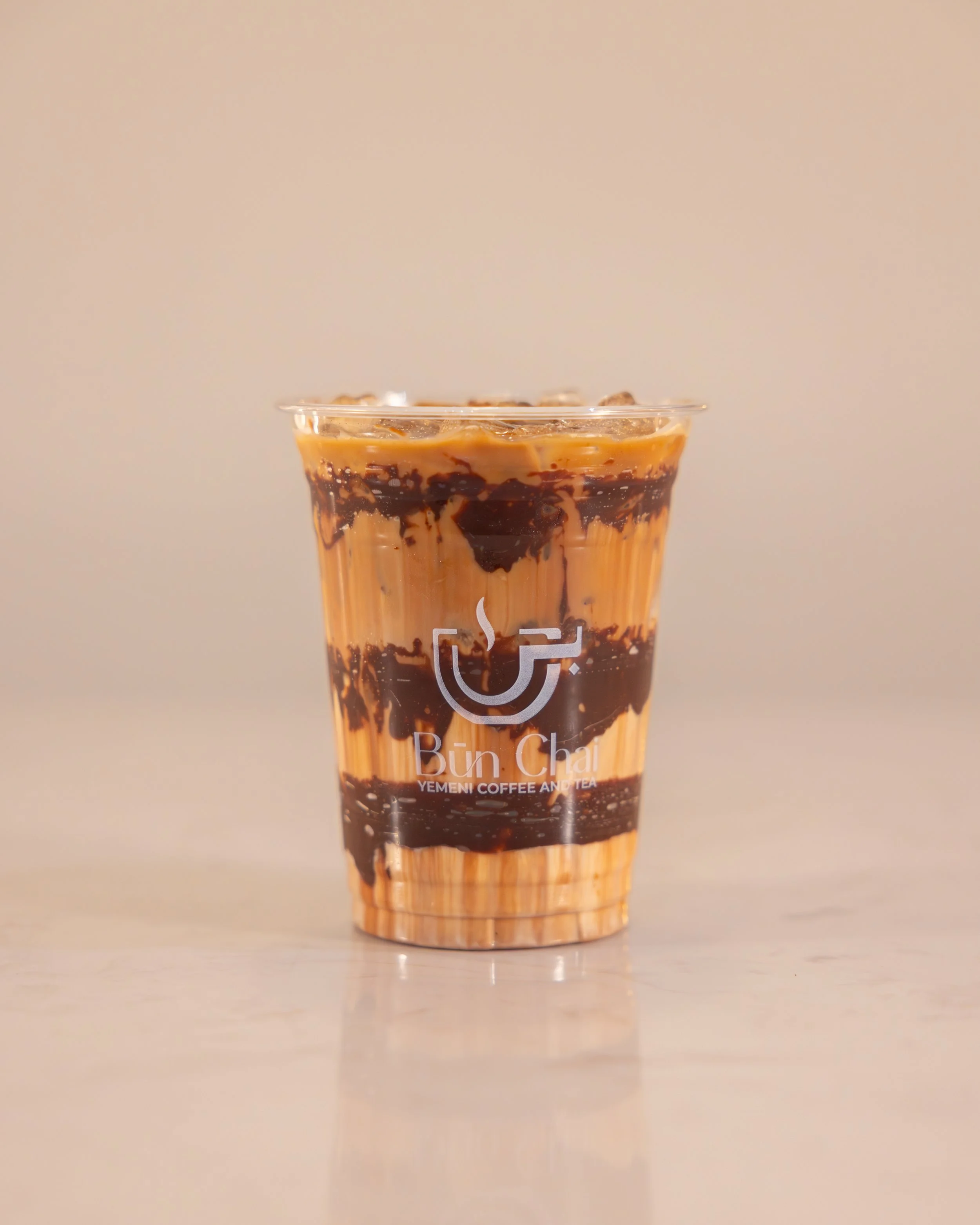 A plastic cup filled with a layered iced coffee drink featuring chocolate syrup and cream, with a Buna Chai logo on the front.