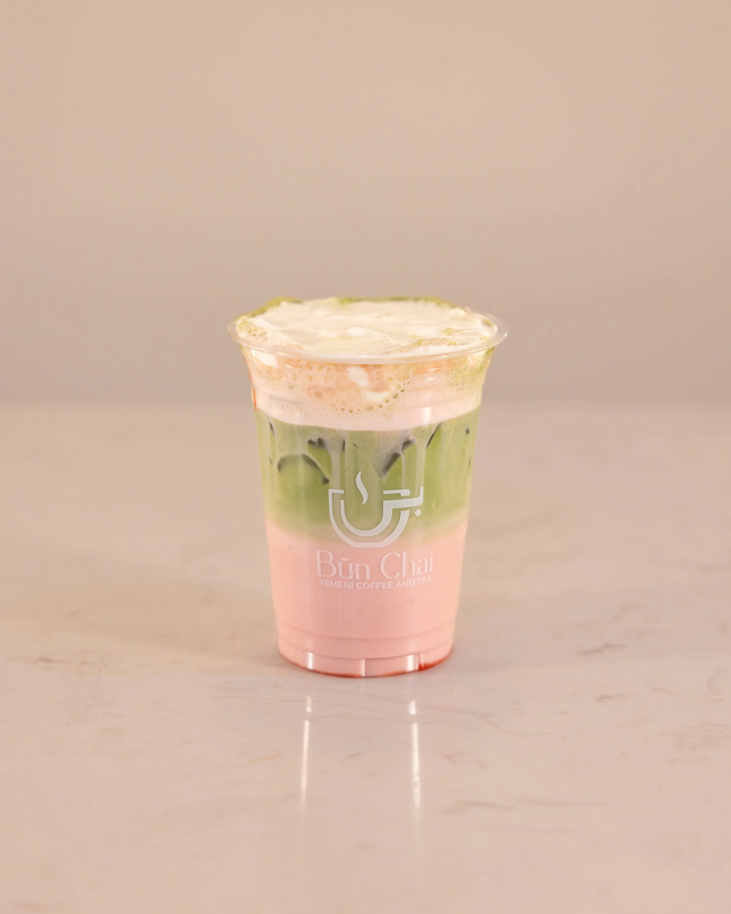 A plastic cup filled with a pink and green layered beverage, topped with foam, placed on a light-colored surface.
