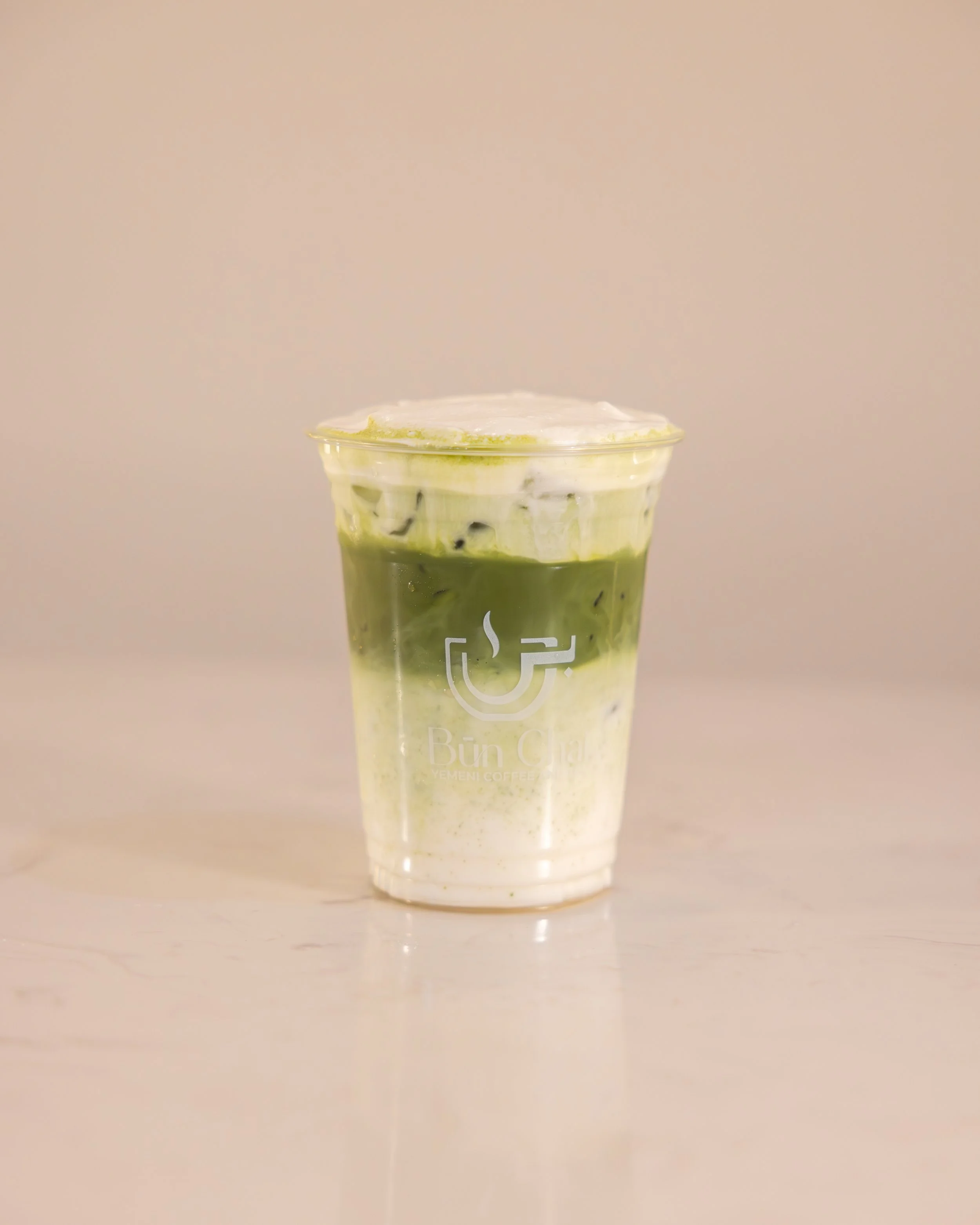 A clear plastic cup filled with iced matcha latte topped with whipped cream, with a logo that says 'Büm Çay' and 'Yemeni Coffee' on the front.