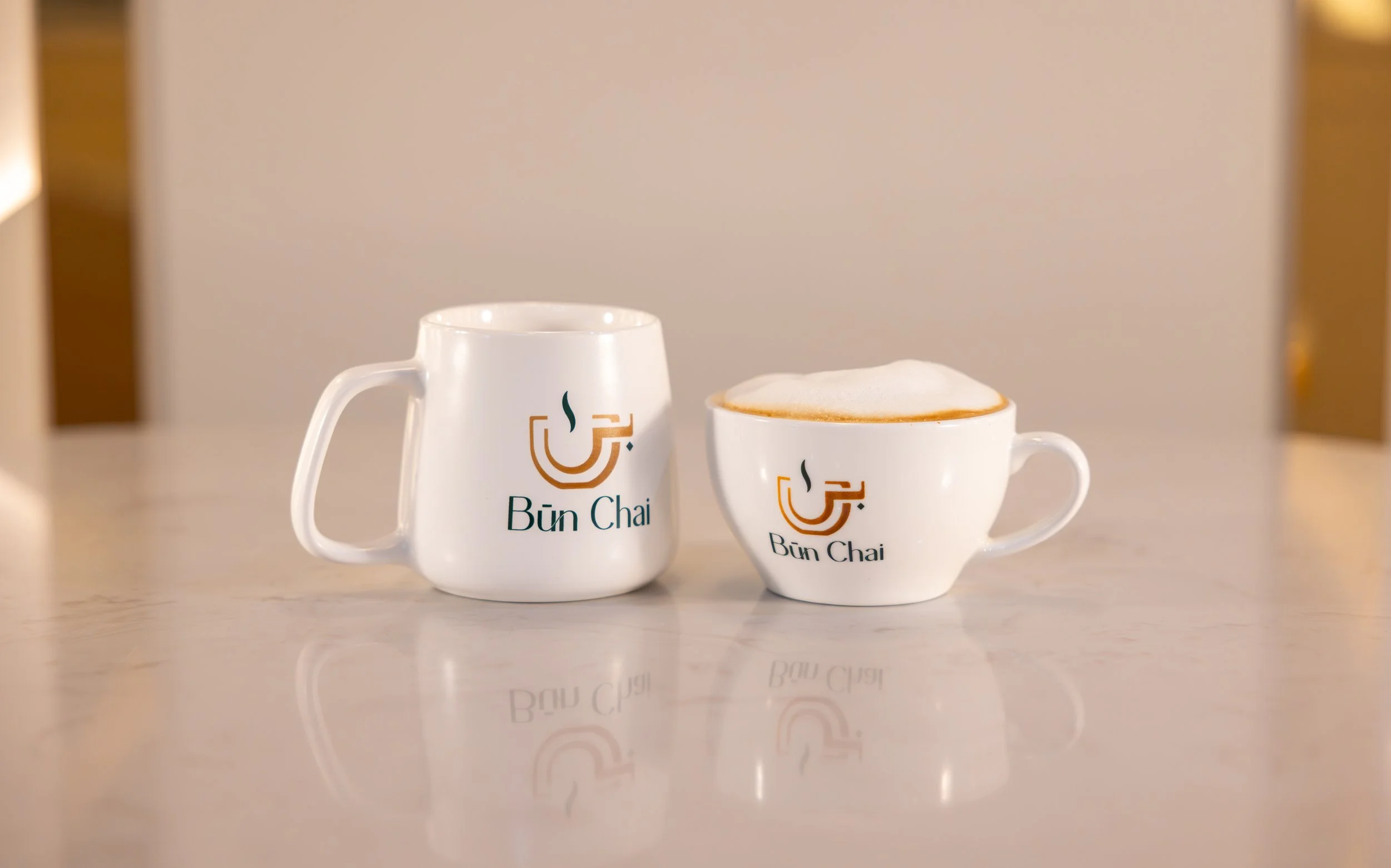 Two white coffee cups with "Bün Chai" logo on a light-colored table, one mug and one cup with latte art foam.