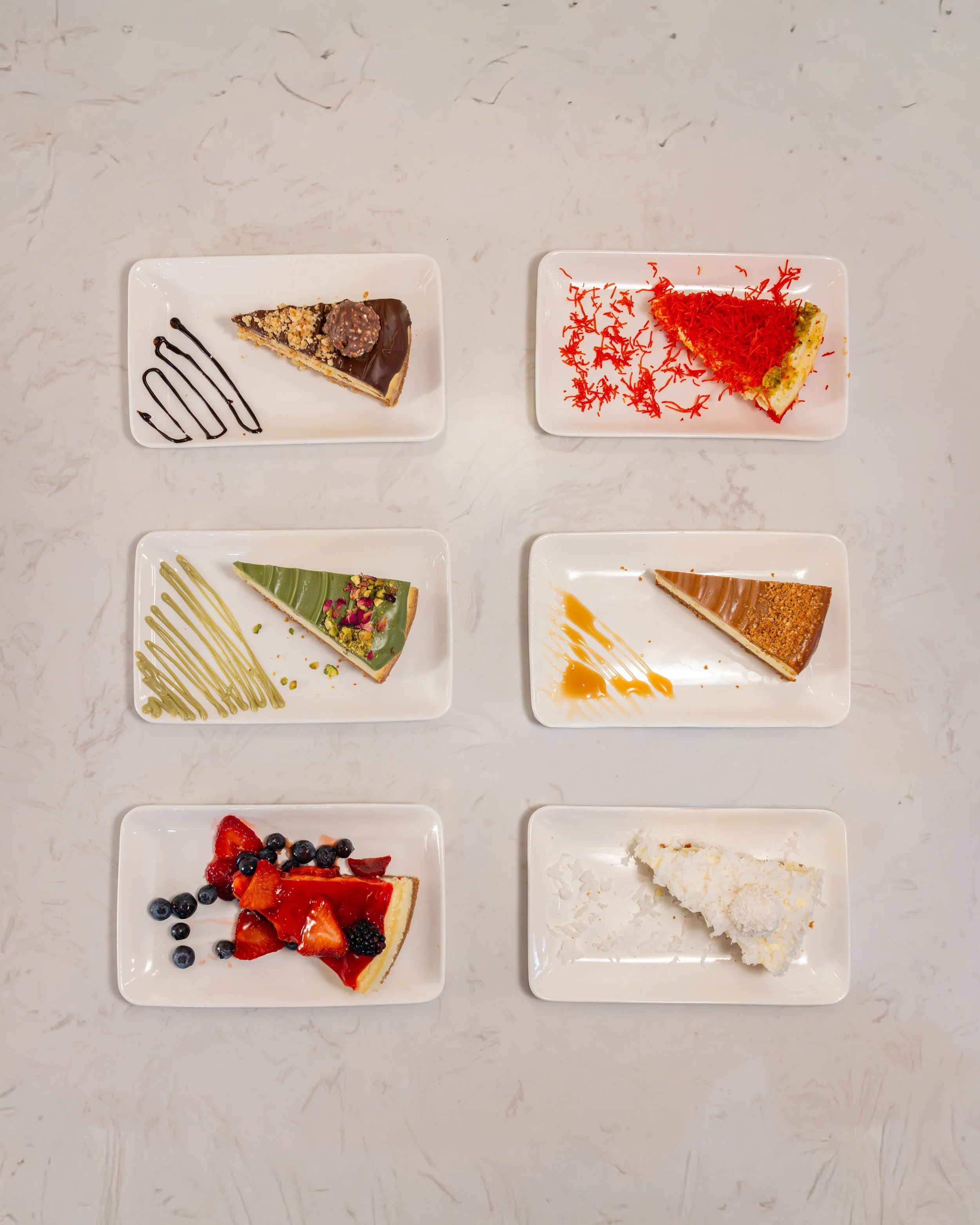 Six plates of assorted desserts arranged on a white marble surface, including a chocolate-topped slice, a red and yellow cake with shredded topping, a green tea and rose pistachio slice, a caramel and hazelnut slice, a berry-topped tart, and a white 