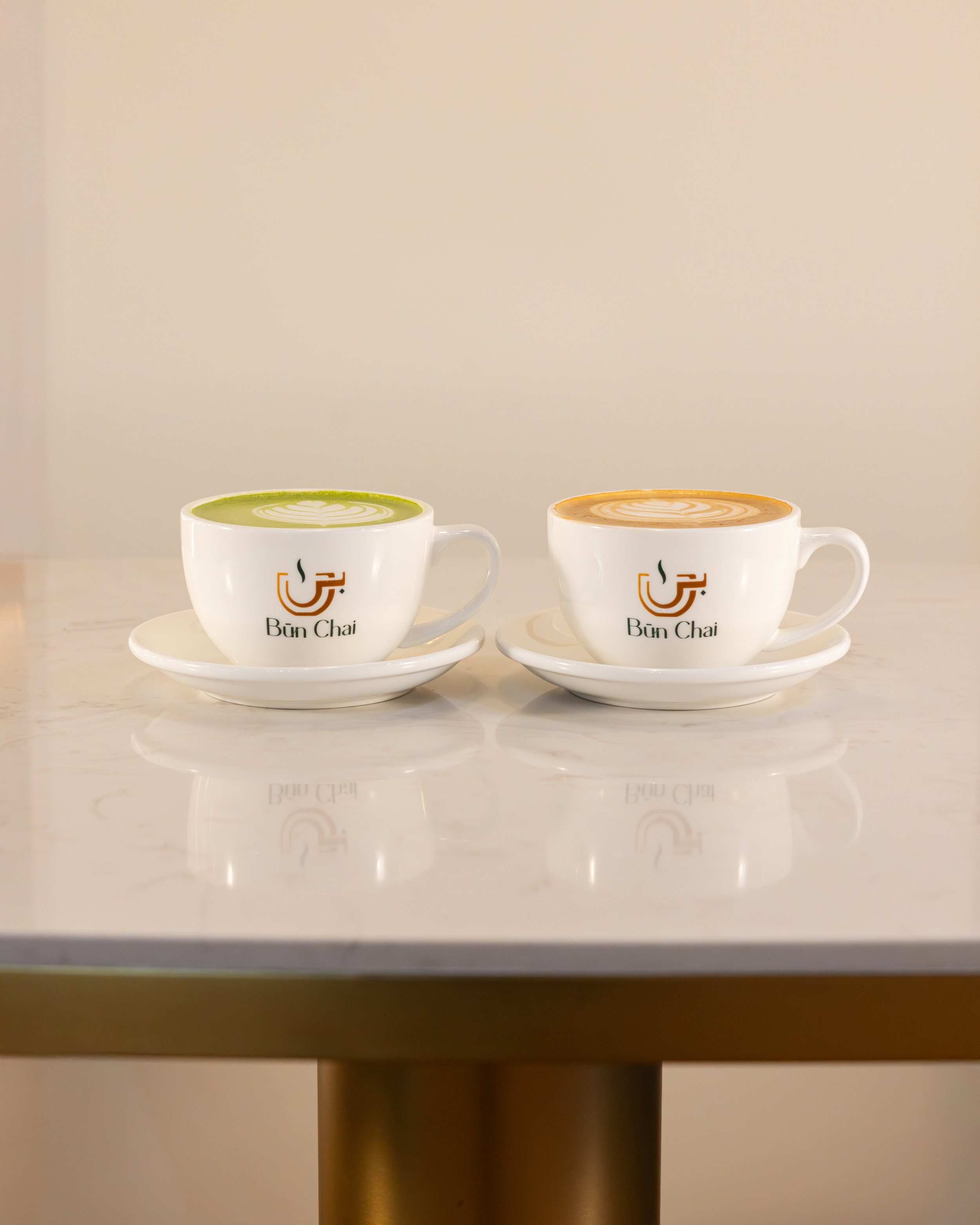 Two white cups with saucers on a marble table, each filled with latte art; one green tea latte and one caramel latte, both with the logo 'Bun Chai' on the cups.