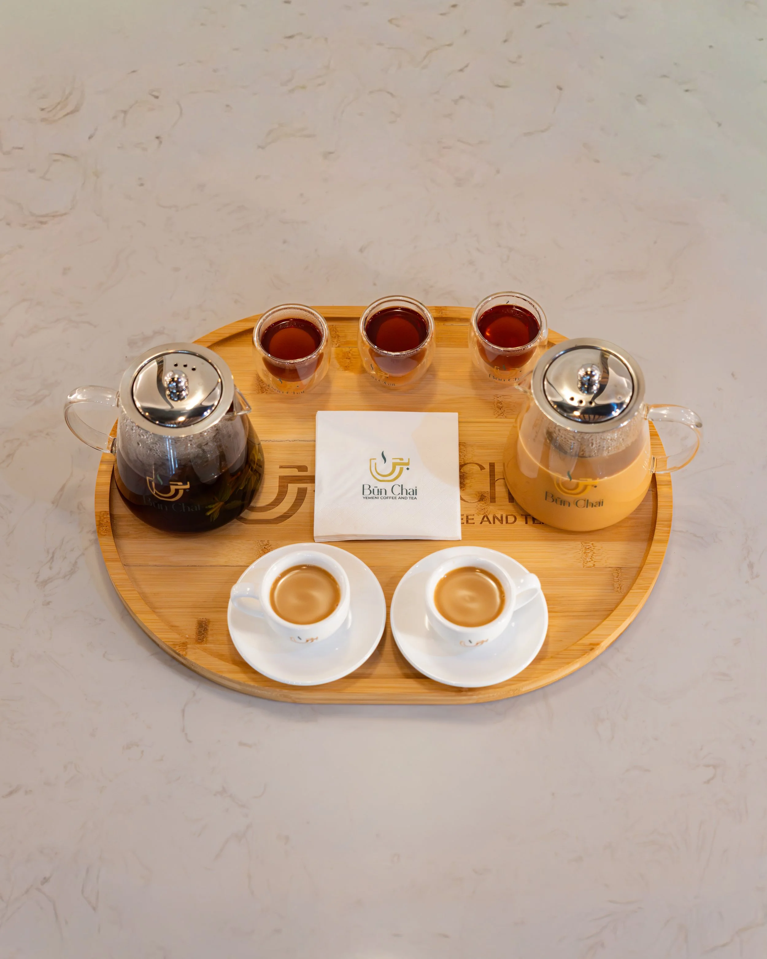 Tray with three small glasses of tea, two cups of coffee with milk, two teapots, and a paper napkin with a logo and text, on a marble surface.
