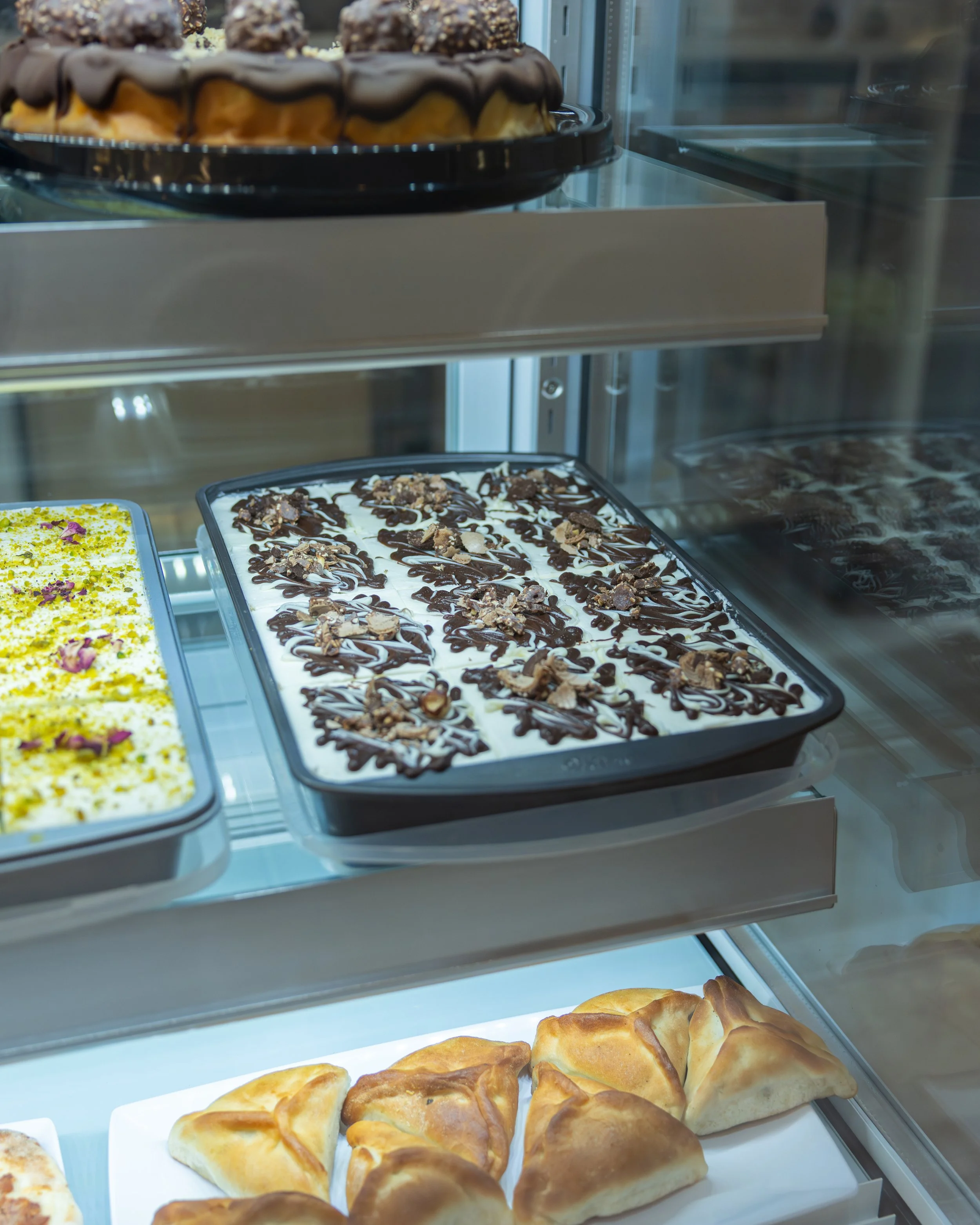 Display case with various desserts including chocolate-drizzled ice cream, a yellow dessert topped with flower petals, and baked pastries.