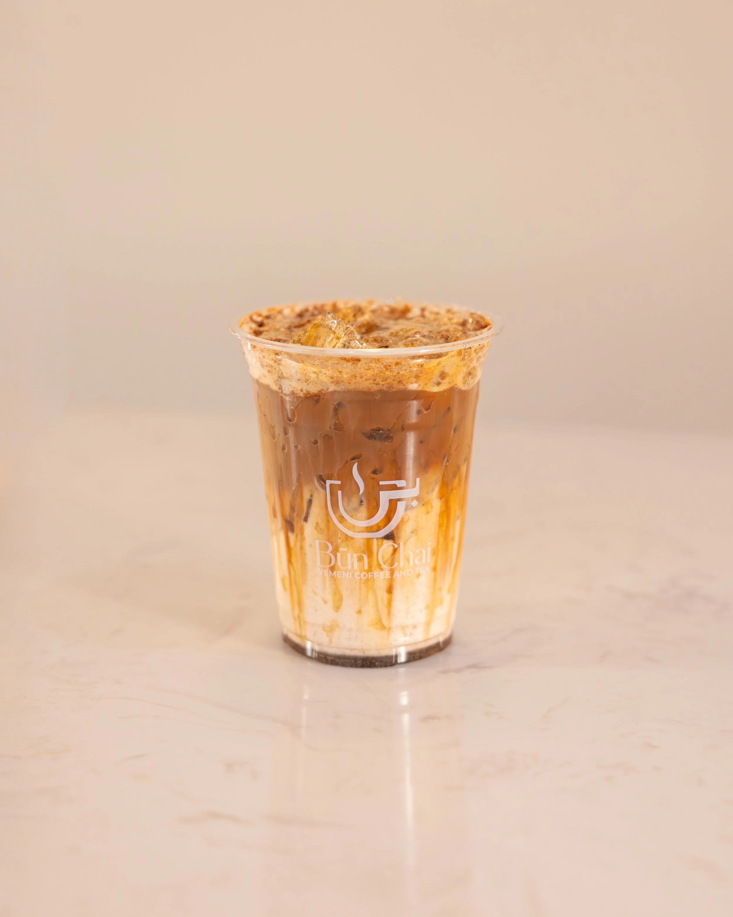 Iced coffee drink in a clear plastic cup with Bun Chai logo, with cream, caramel, and chocolate syrup, served on a white surface.