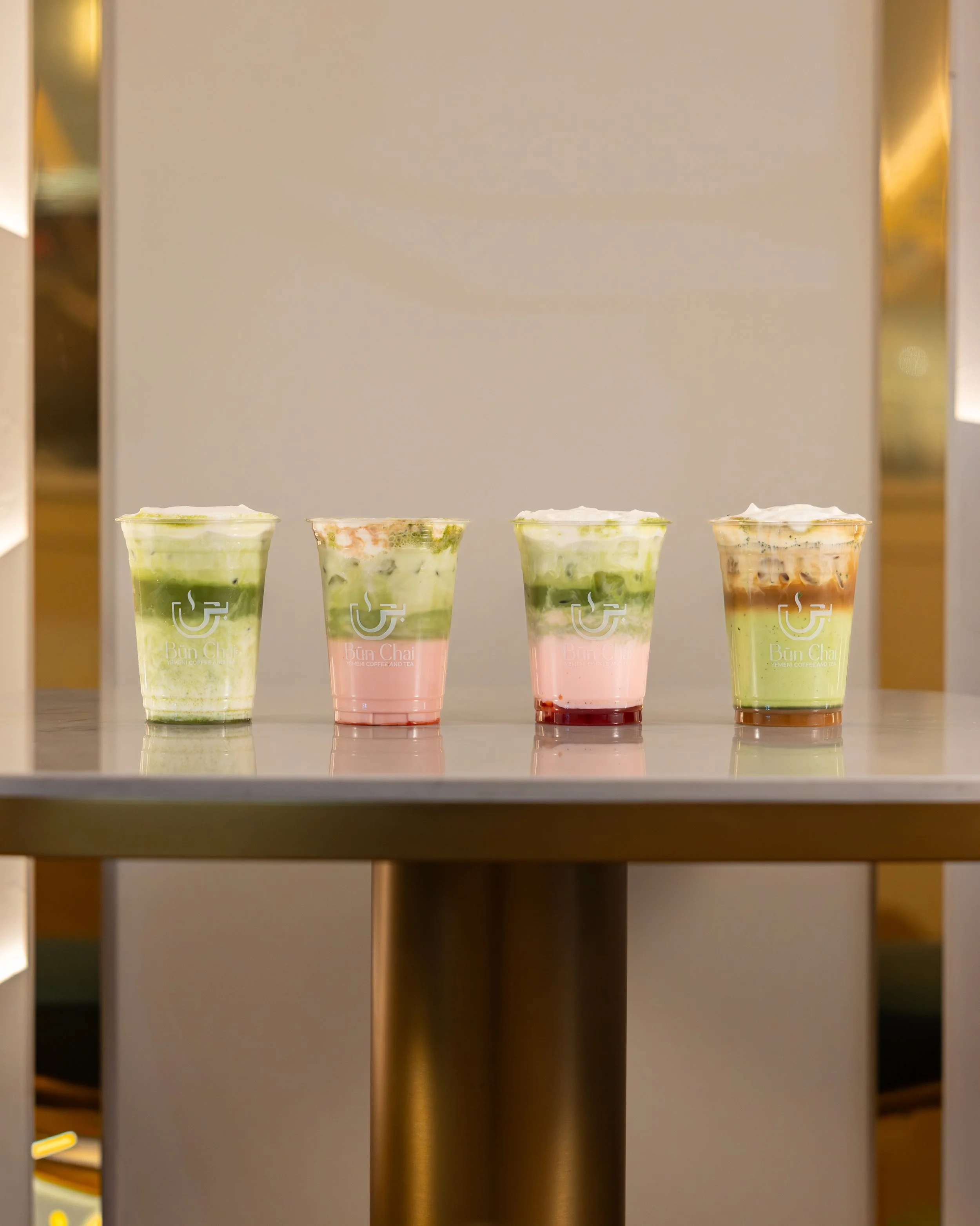 Four colorful beverages in plastic cups on a table, with a neutral background. Each drink has different layers and toppings.