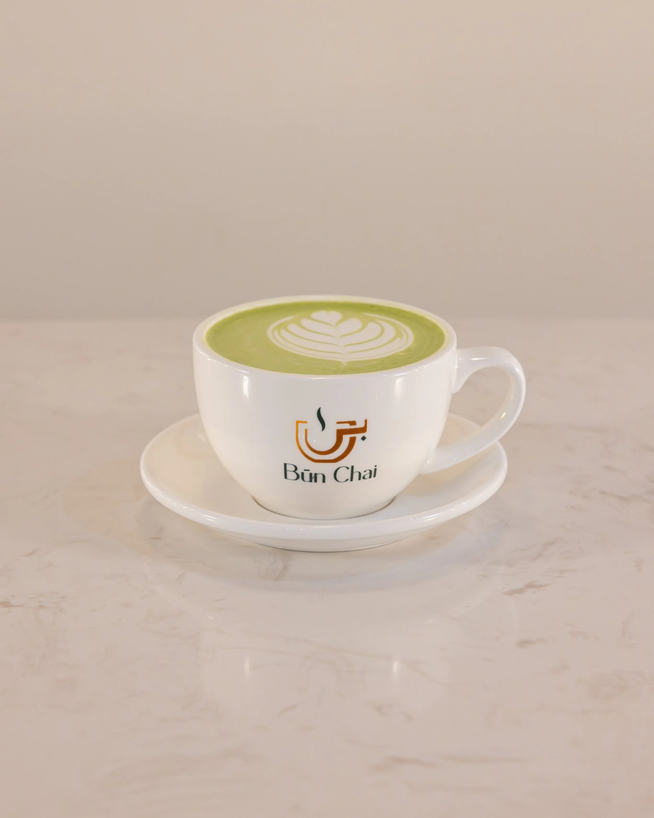 A white cup of matcha latte with latte art on top, sitting on a white saucer, with 'Bun Chai' logo on the cup.