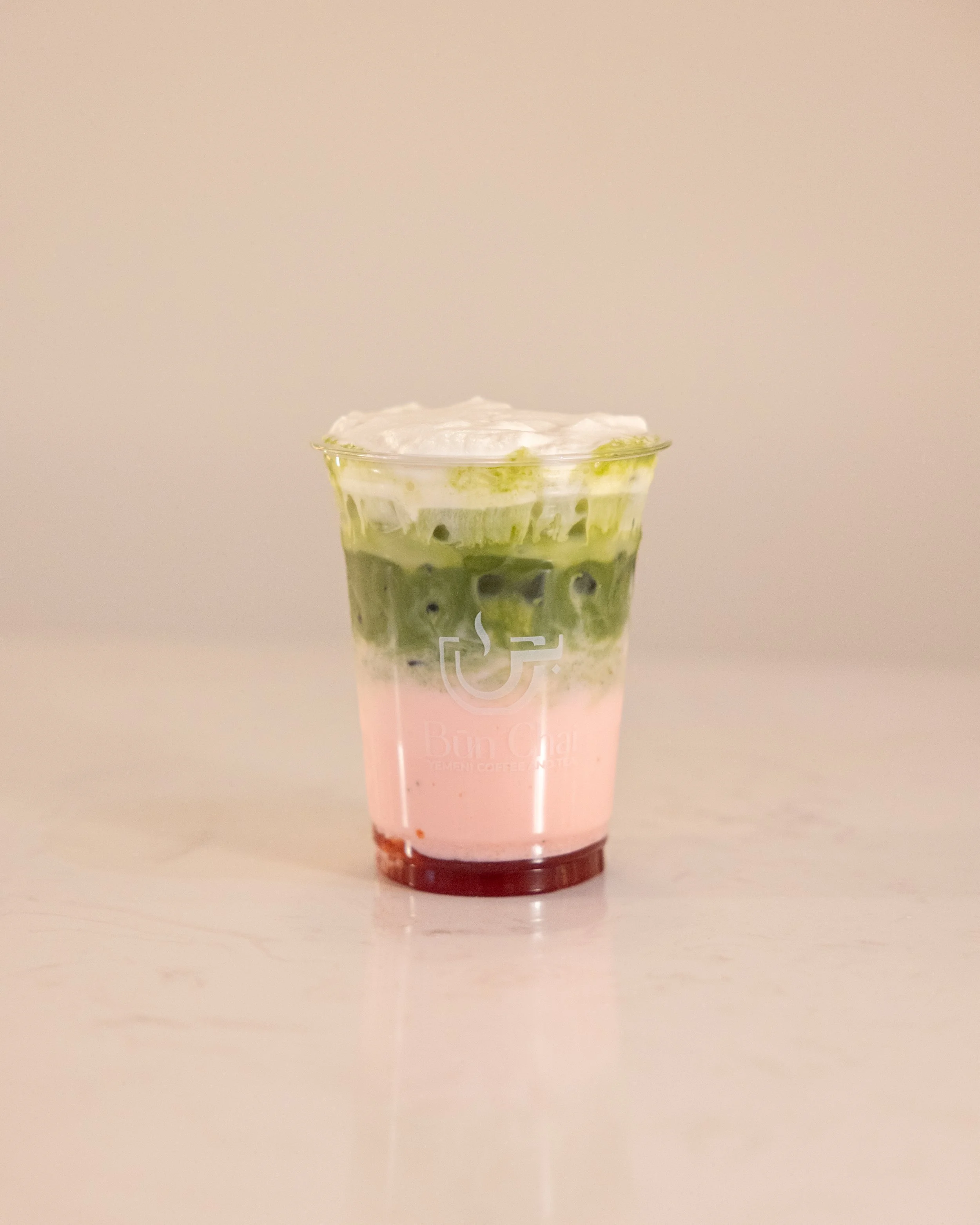 Layered drink with pink, green, and white layers, topped with whipped cream, in a clear plastic cup from Bun Cha.