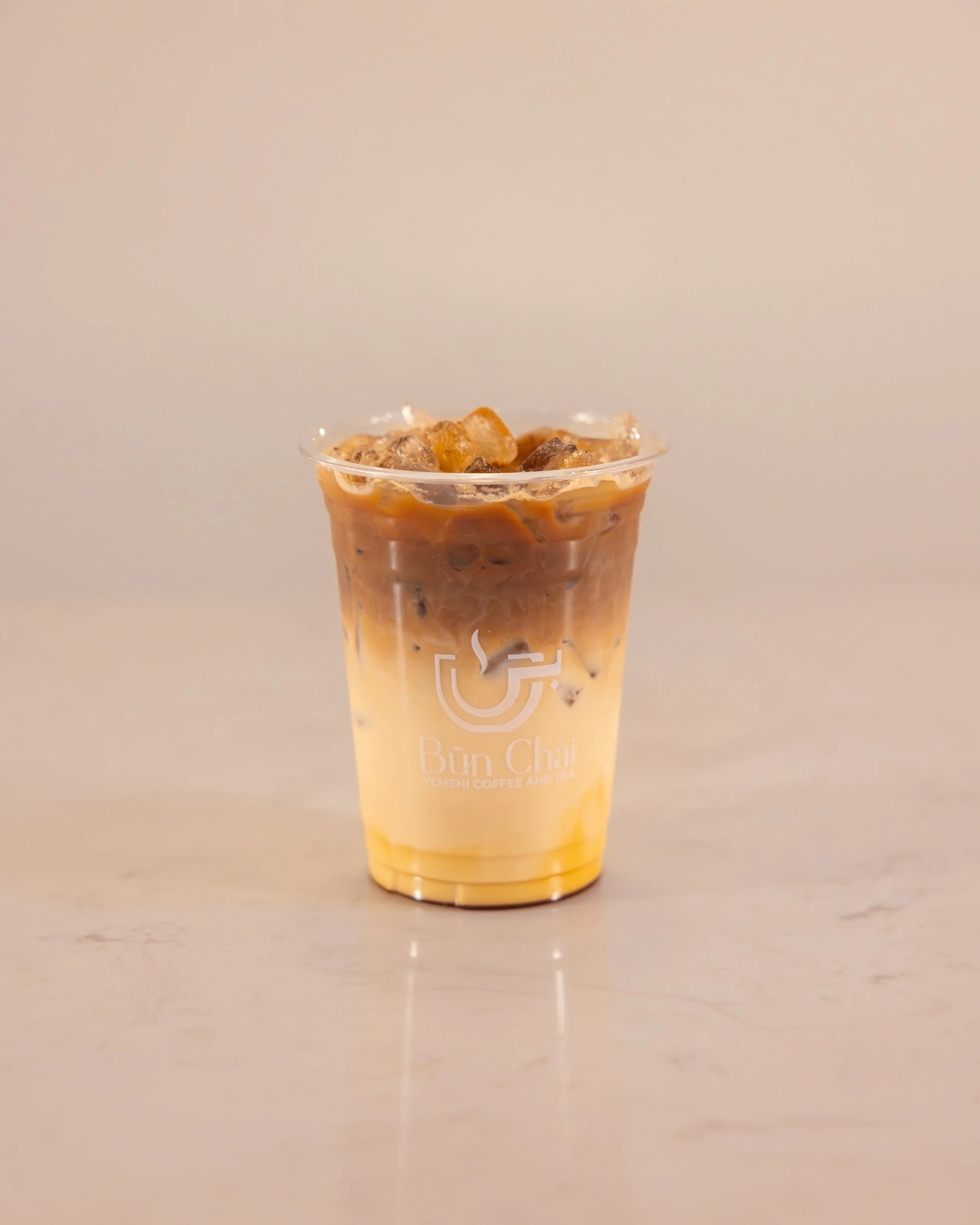 Ice coffee drink in a clear plastic cup with a logo, containing ice cubes and layered coffee and milk.