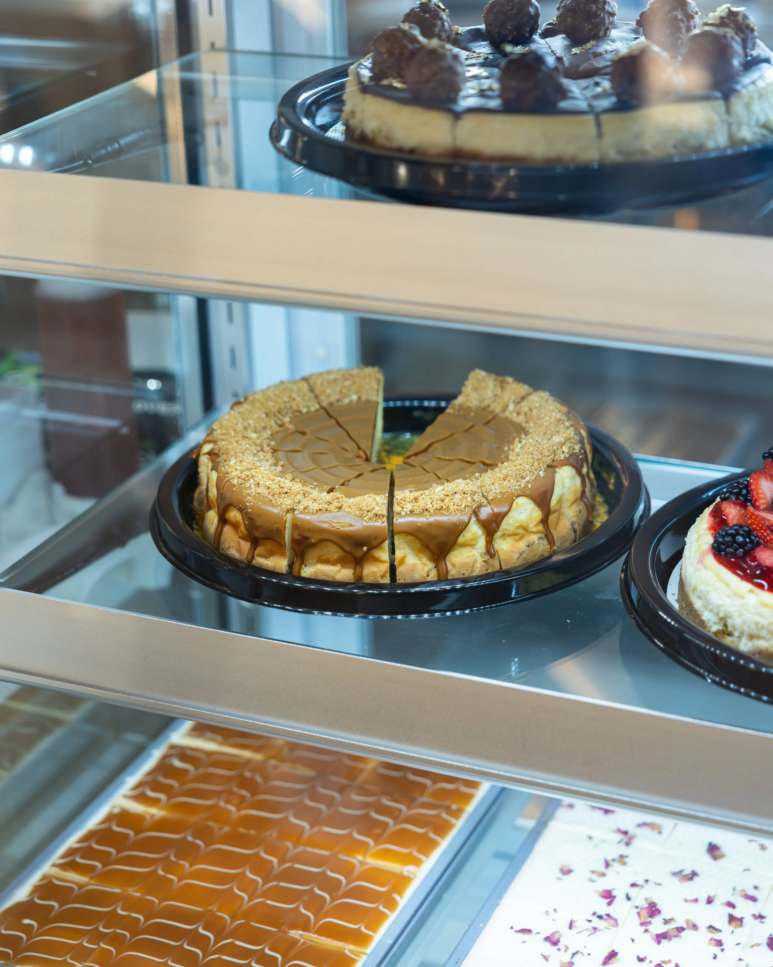 Display case with various cakes, including a caramel-flavored cheesecake topped with chocolate, nuts, and caramel drizzle, and a strawberry-topped cheesecake with berry garnish.
