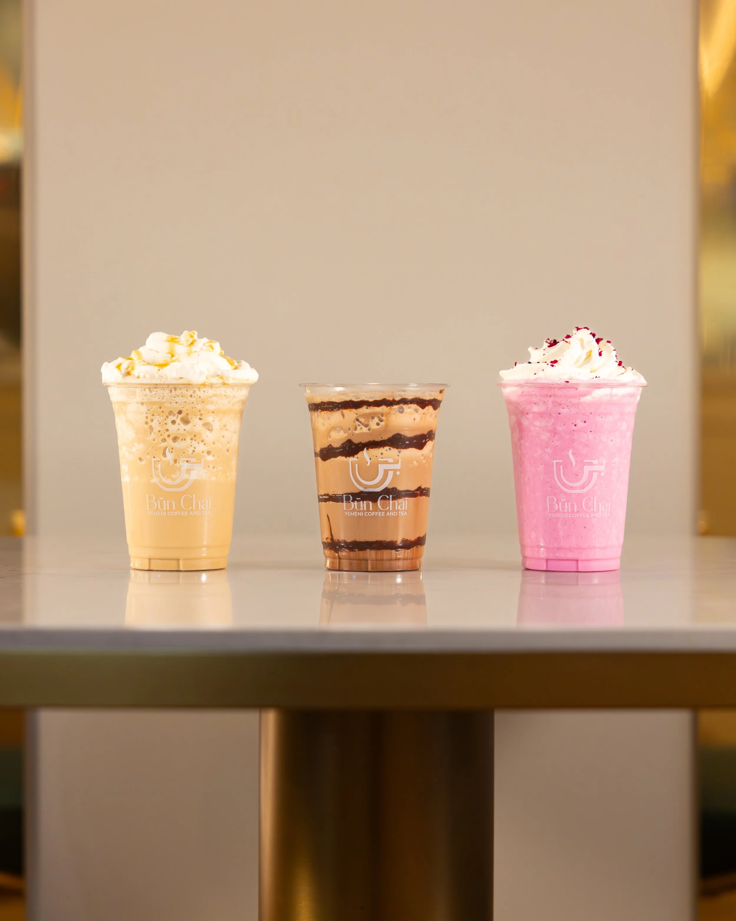Three colorful beverages on a table; on the left a creamy yellow drink topped with whipped cream, in the middle a layered brown and tan drink with chocolate syrup, and on the right a pink drink topped with whipped cream and red sprinkles, all in clea