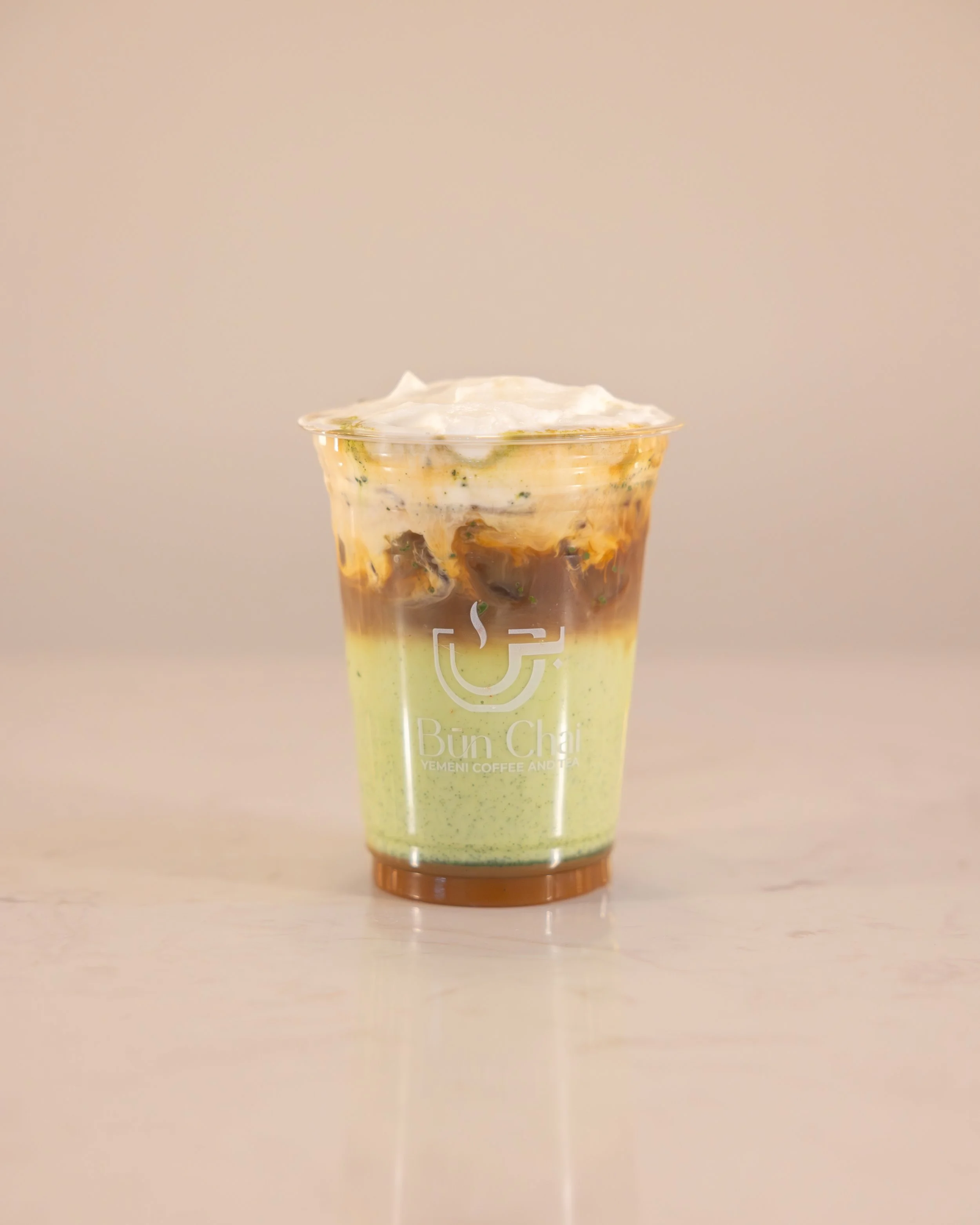 A layered iced beverage with green, brown, and white layers in a clear cup with a logo, topped with whipped cream.