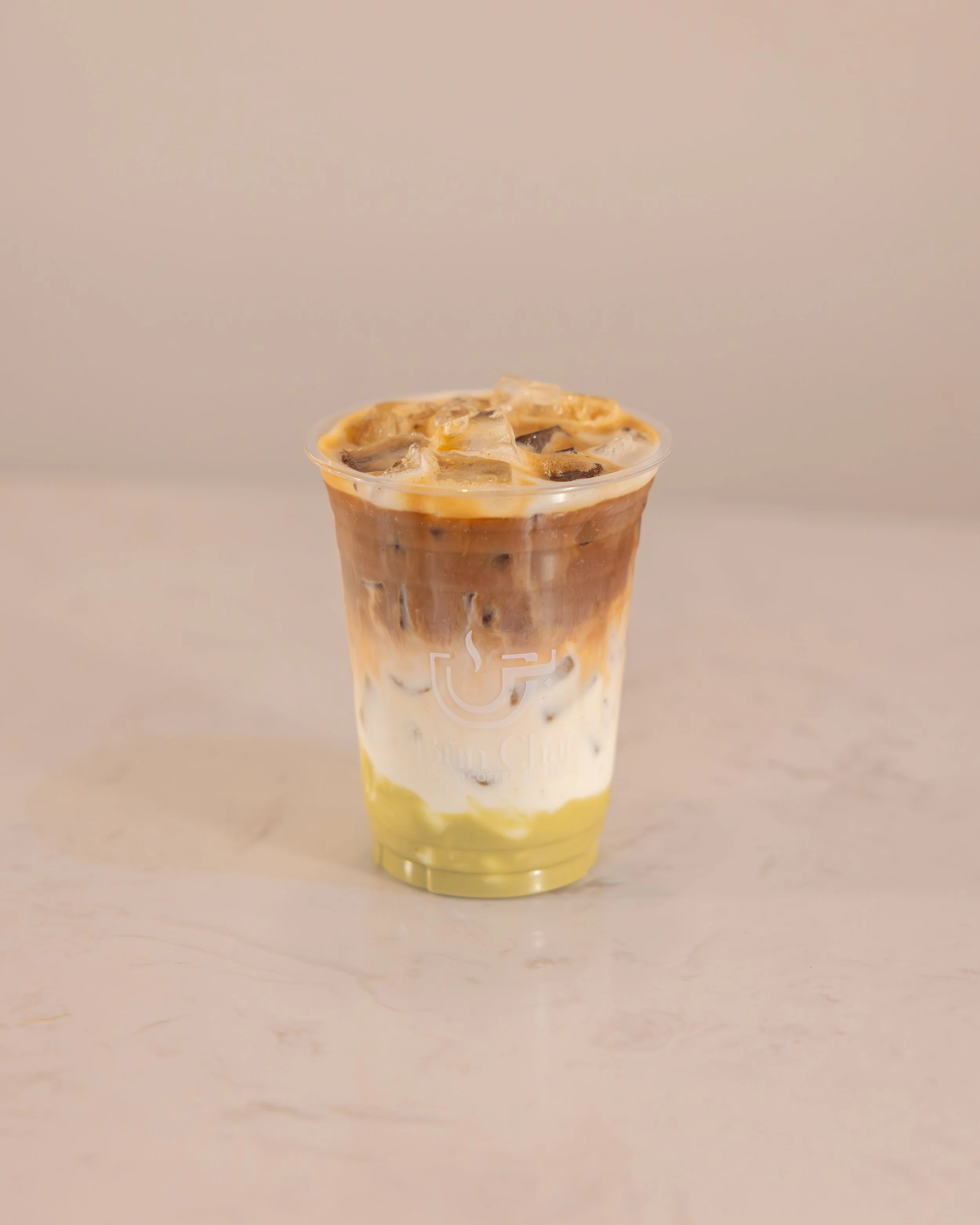 A clear plastic cup filled with iced coffee layered with milk and green matcha at the bottom, with ice cubes on top.