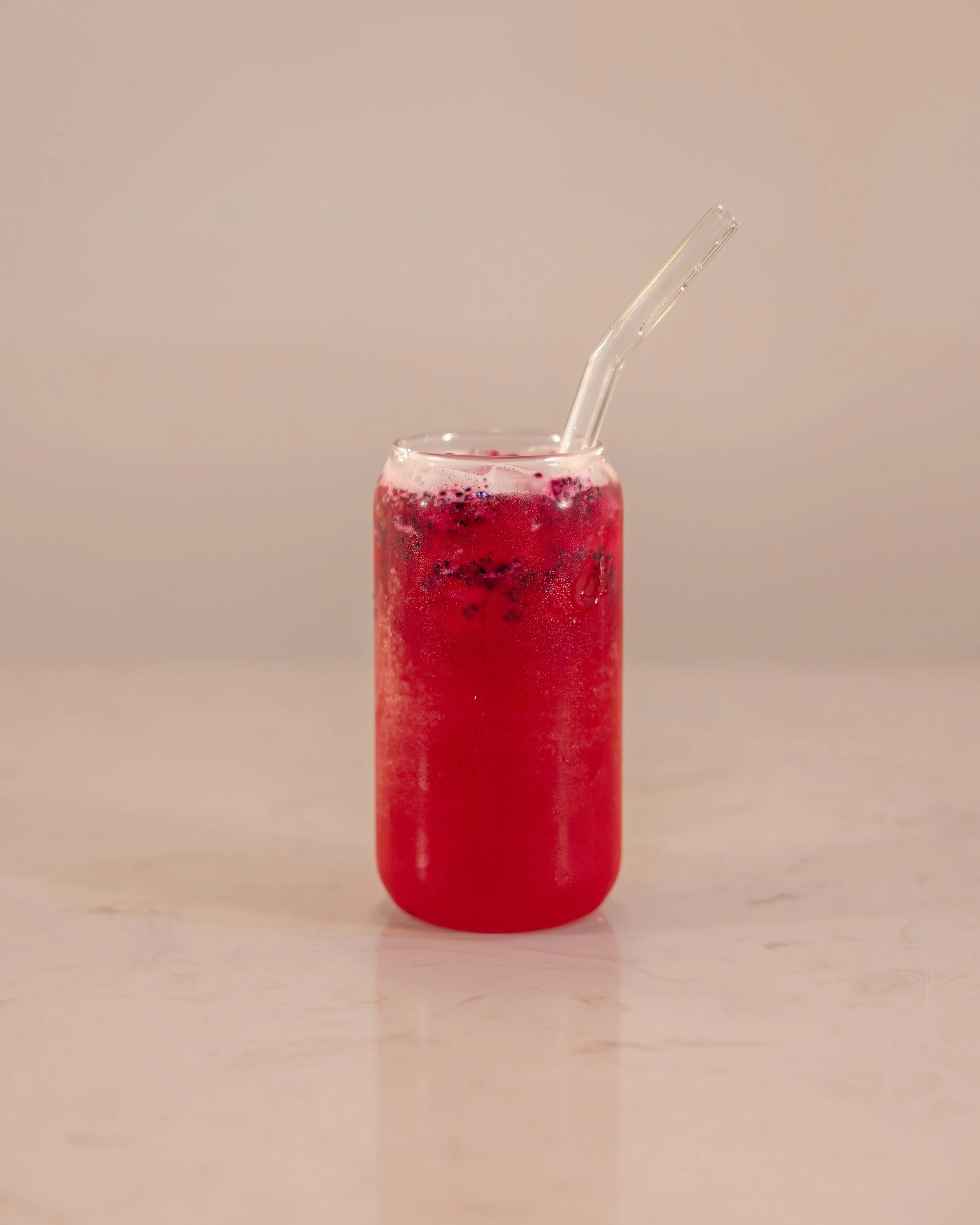 A tall glass of red berry soda with condensation, a clear plastic straw, and bubbles visible inside, on a light-colored surface against a plain background.