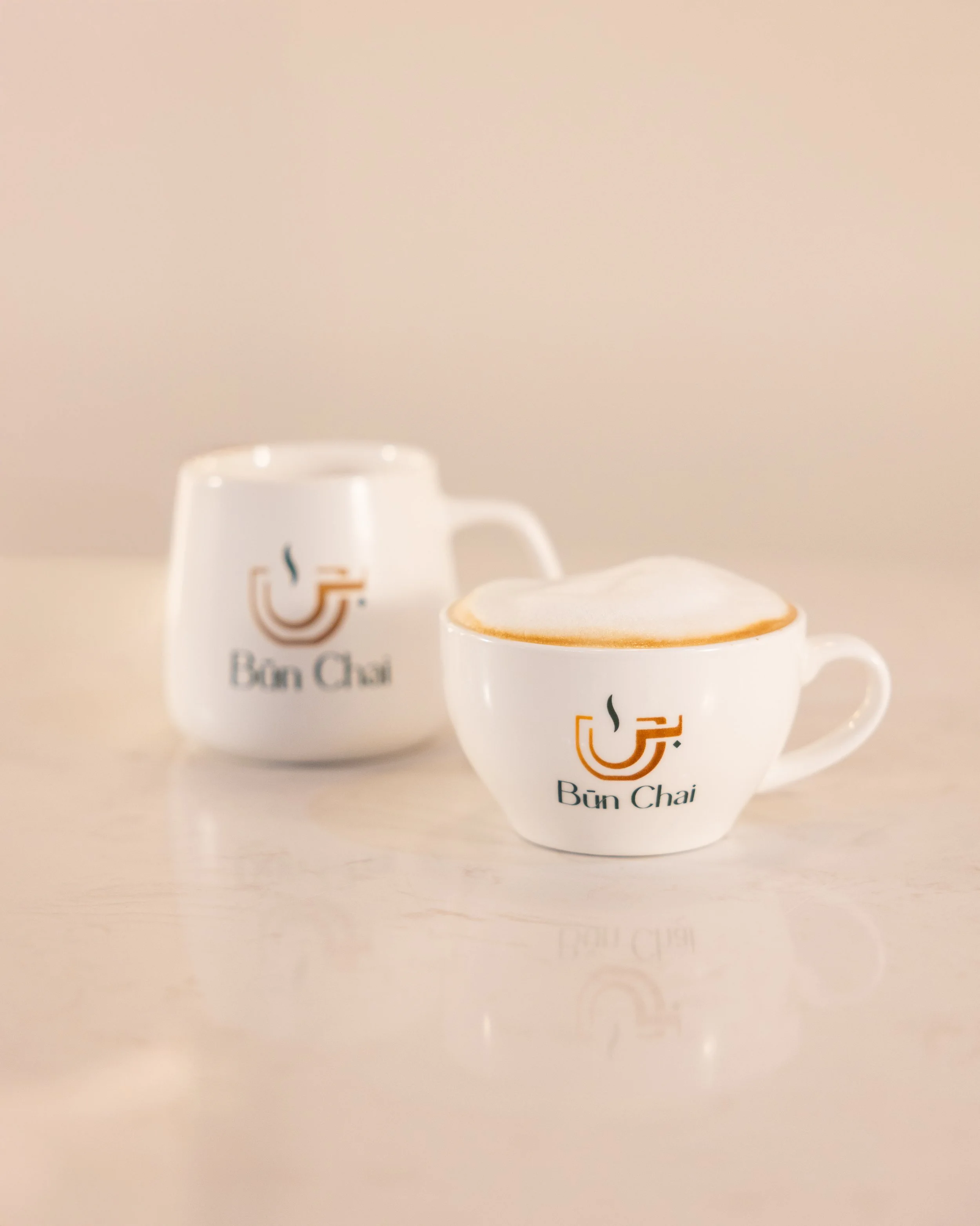 Two white coffee cups with the Bun Chai logo, one filled with a frothy cappuccino and the other empty, on a light-colored surface.