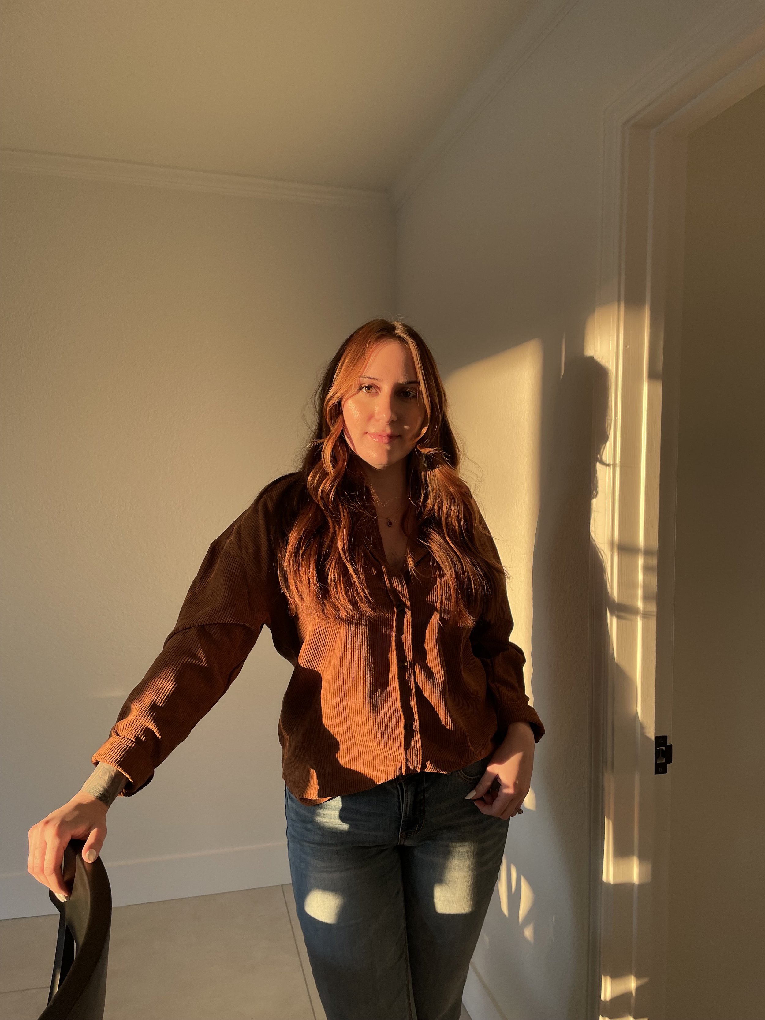 A woman with long brown hair stands in a warmly lit room, casting a shadow on the wall behind her. She is wearing a brown shirt and blue jeans, with sunlight illuminating her face and upper body.