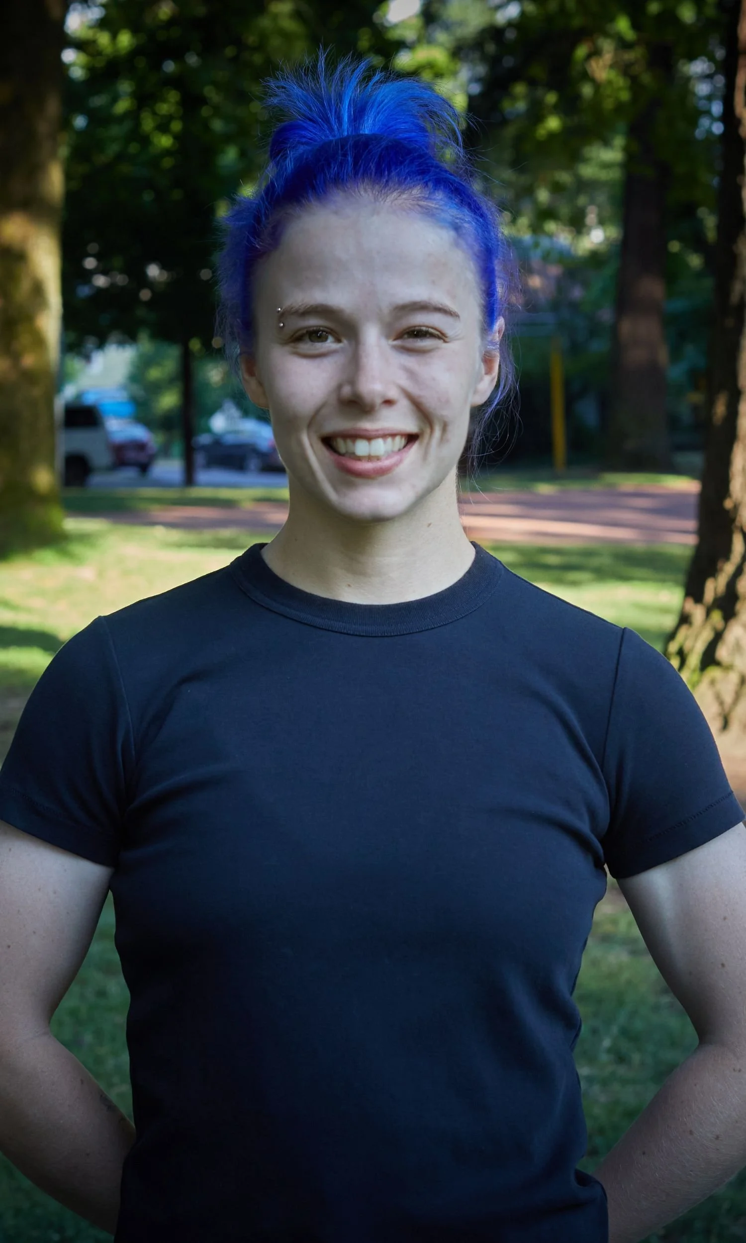 Owner and Coach, Megan, has bright purple hair styled in a messy bun, smiling outdoors in a park.