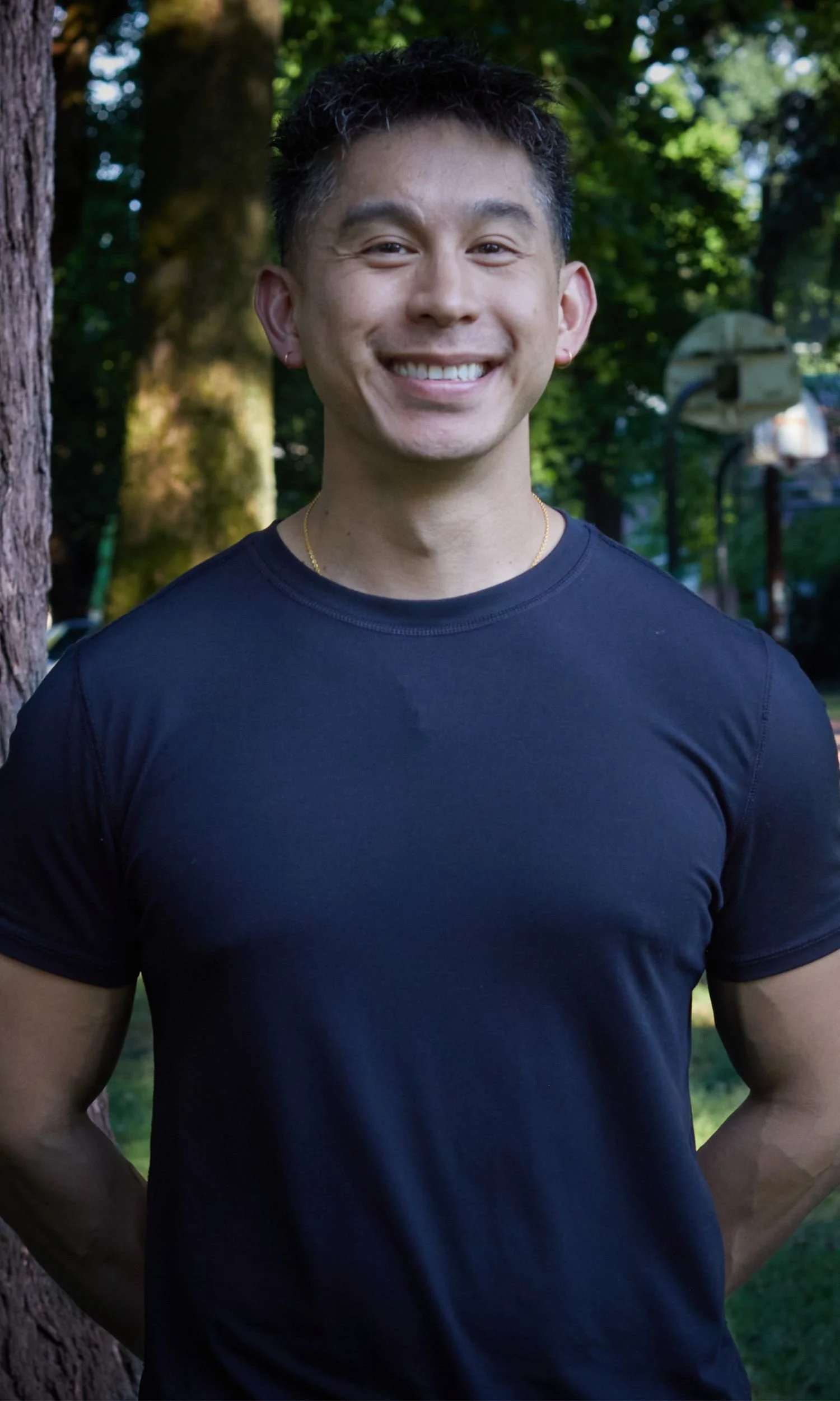 Owner and Coach, Joe, close-up headshot smiling man with short black hair, wearing a black T-shirt, outdoors in a park with green trees and sunlight.