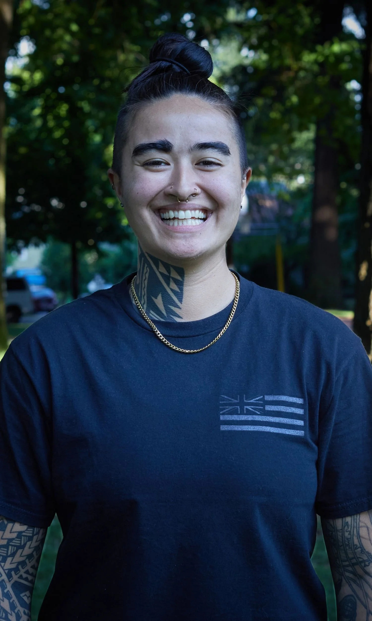 Owner and Coach, Chase, a woman with a bun hairstyle, smiling in a park with green trees, wearing a black T-shirt with a Hawaii flag and tattooed arms.