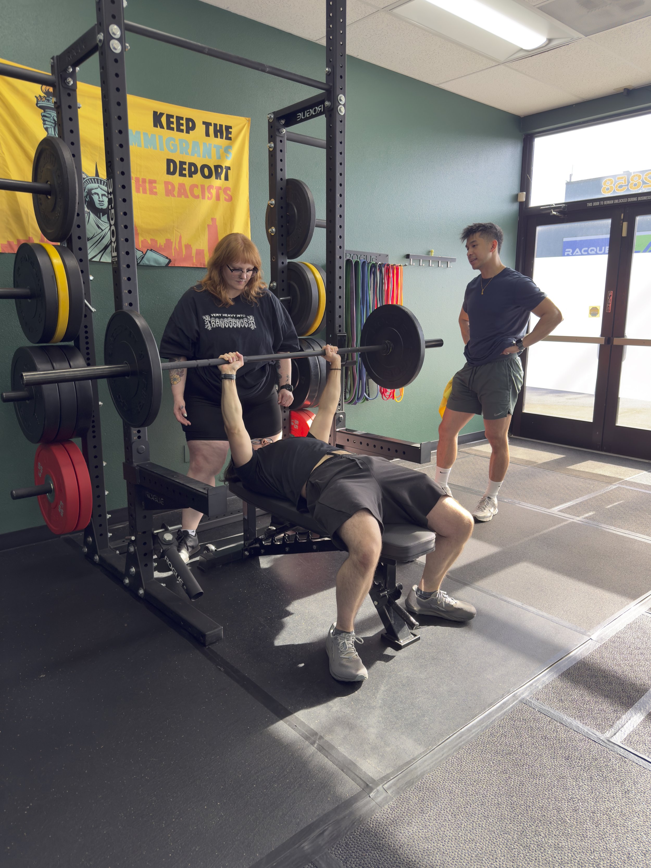 A woman spotting on a bench press with a barbell in a gym, Coach Joe standing nearby for safety. Resistance bands hanging on the wall.