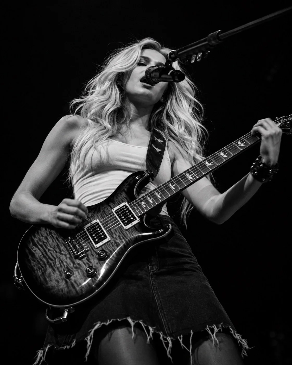 A woman with long wavy blonde hair playing an electric guitar and singing into a microphone on stage